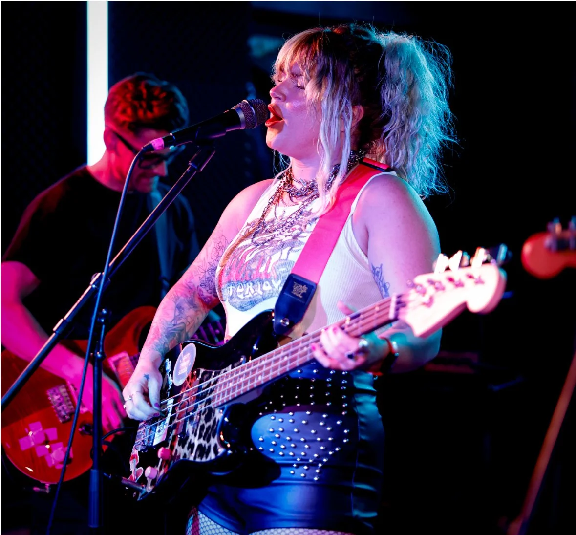 Female musician with blonde, curly hair singing into a microphone while playing an electric guitar on stage, with a male guitarist in the background. Singing Lessons Bideford, Music Teacher Bideford, Singing Lessons North Devon, Bold Note Vocal Coach