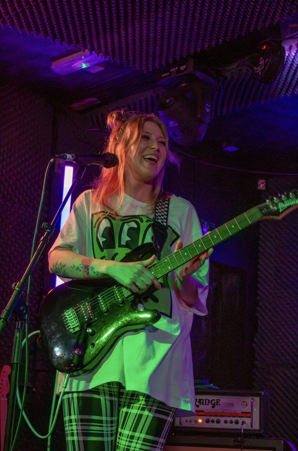 A young woman with red hair playing an electric guitar and smiling on stage under colorful lights.