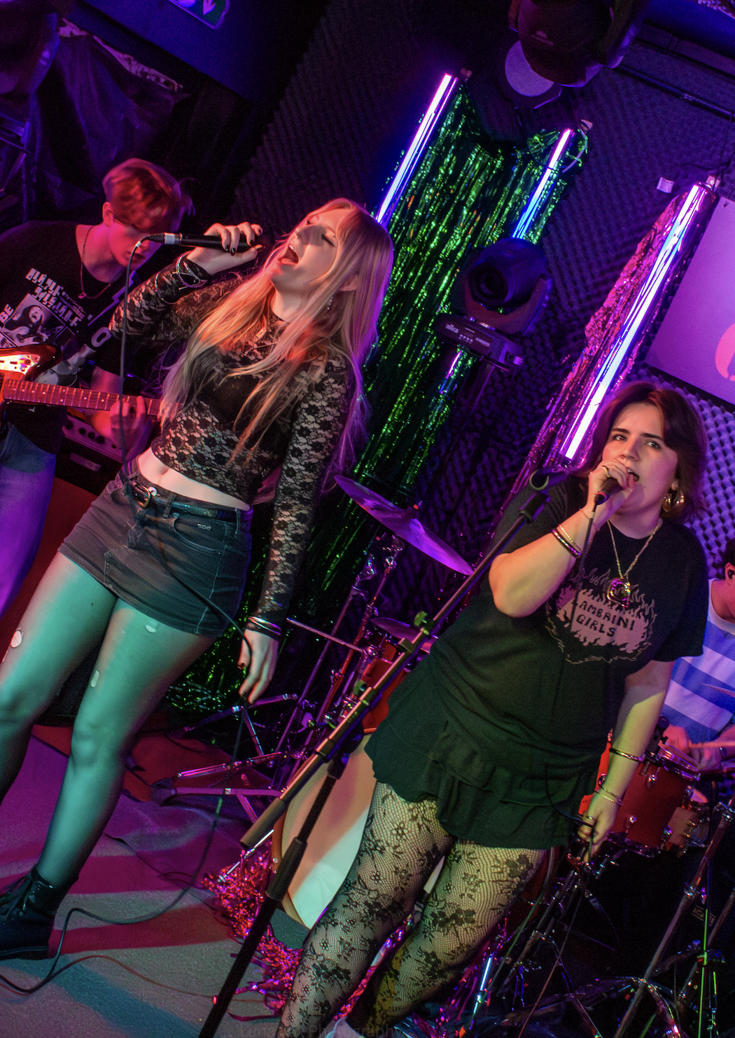 Two female singers perform with band members playing electric guitar and drums in a colorful, neon-lit stage setting.