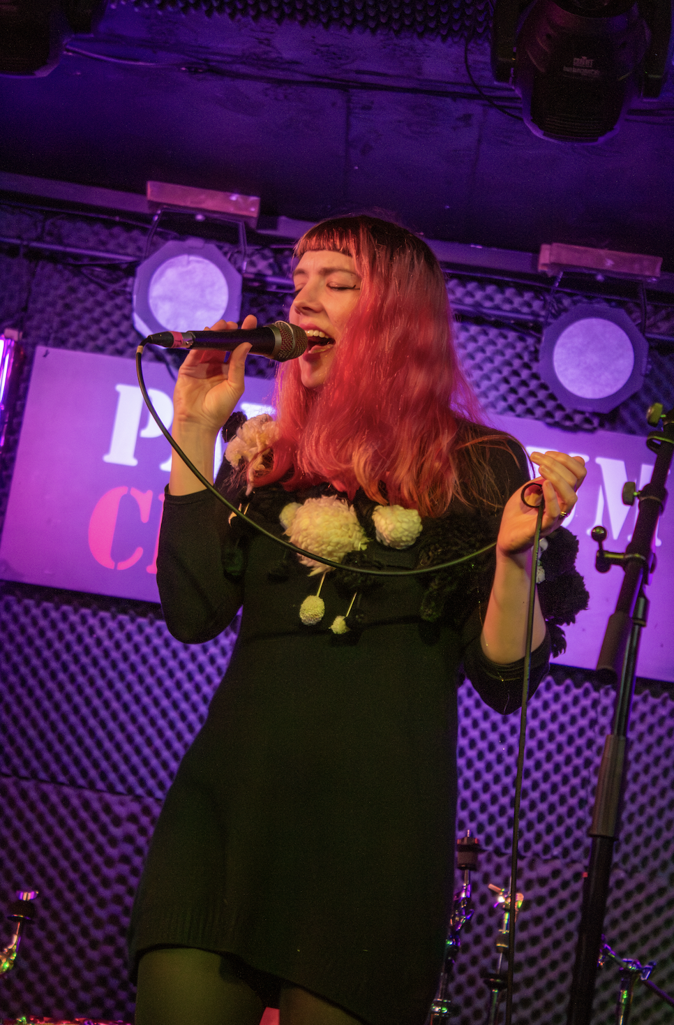 A woman singing into a microphone on stage with purple lighting and a patterned backdrop.