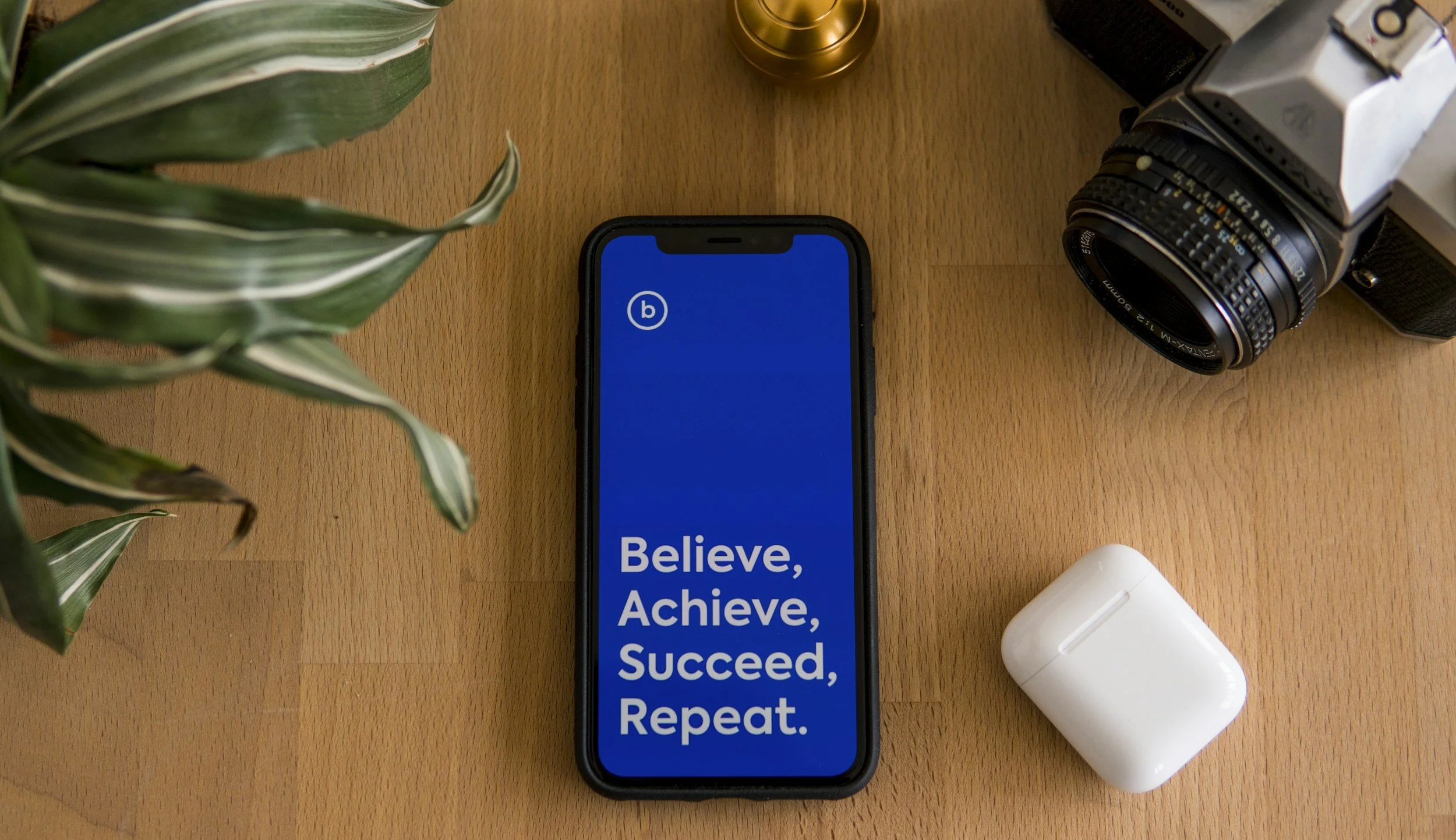 Smartphone with a motivational message, a vintage camera, white wireless earbuds in case, a green leafy plant, and a small gold container on a wooden surface.