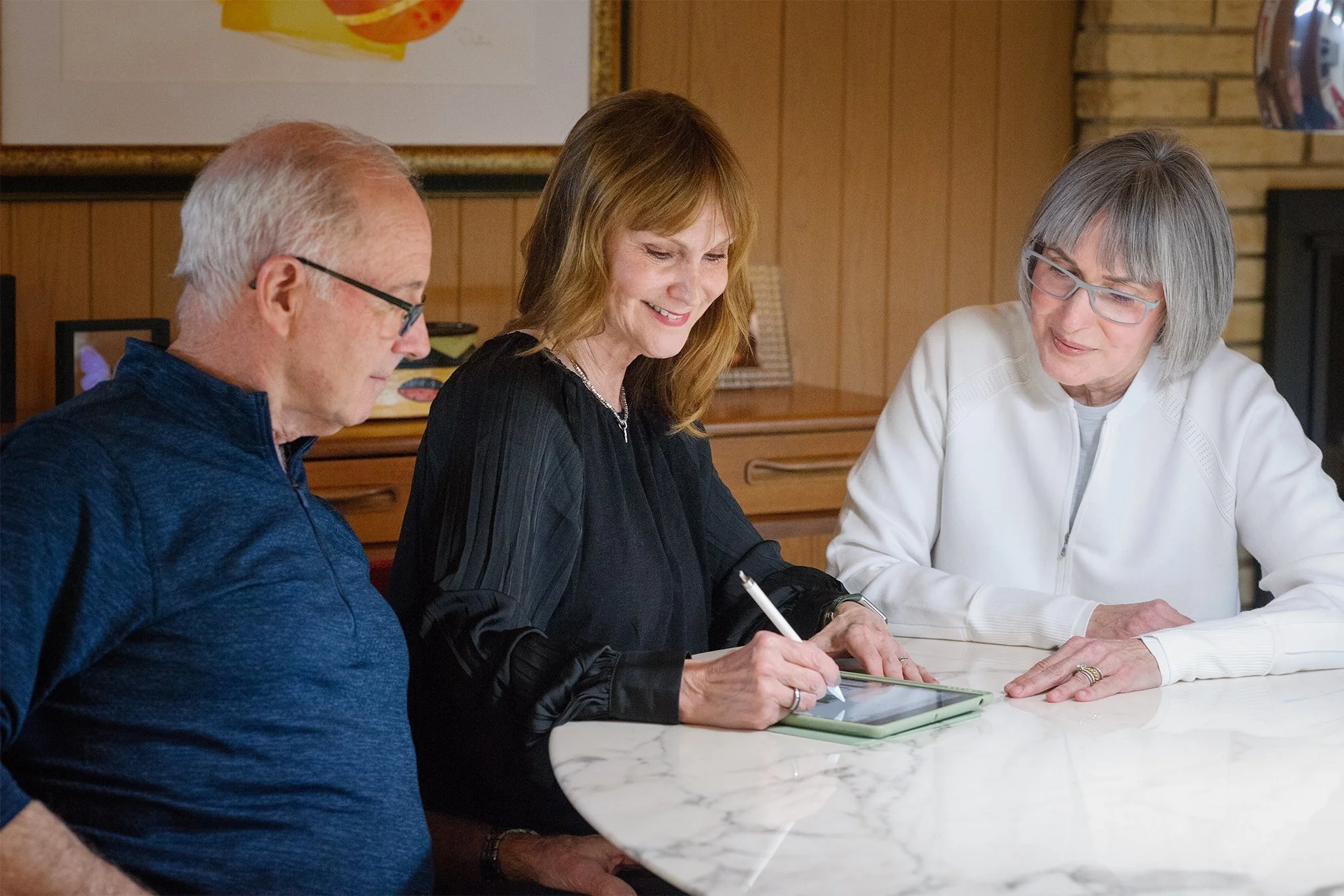 Three older adults, two women and one man, sitting at a white marble table in a cozy room, with one woman using a tablet while the other woman and man look on.