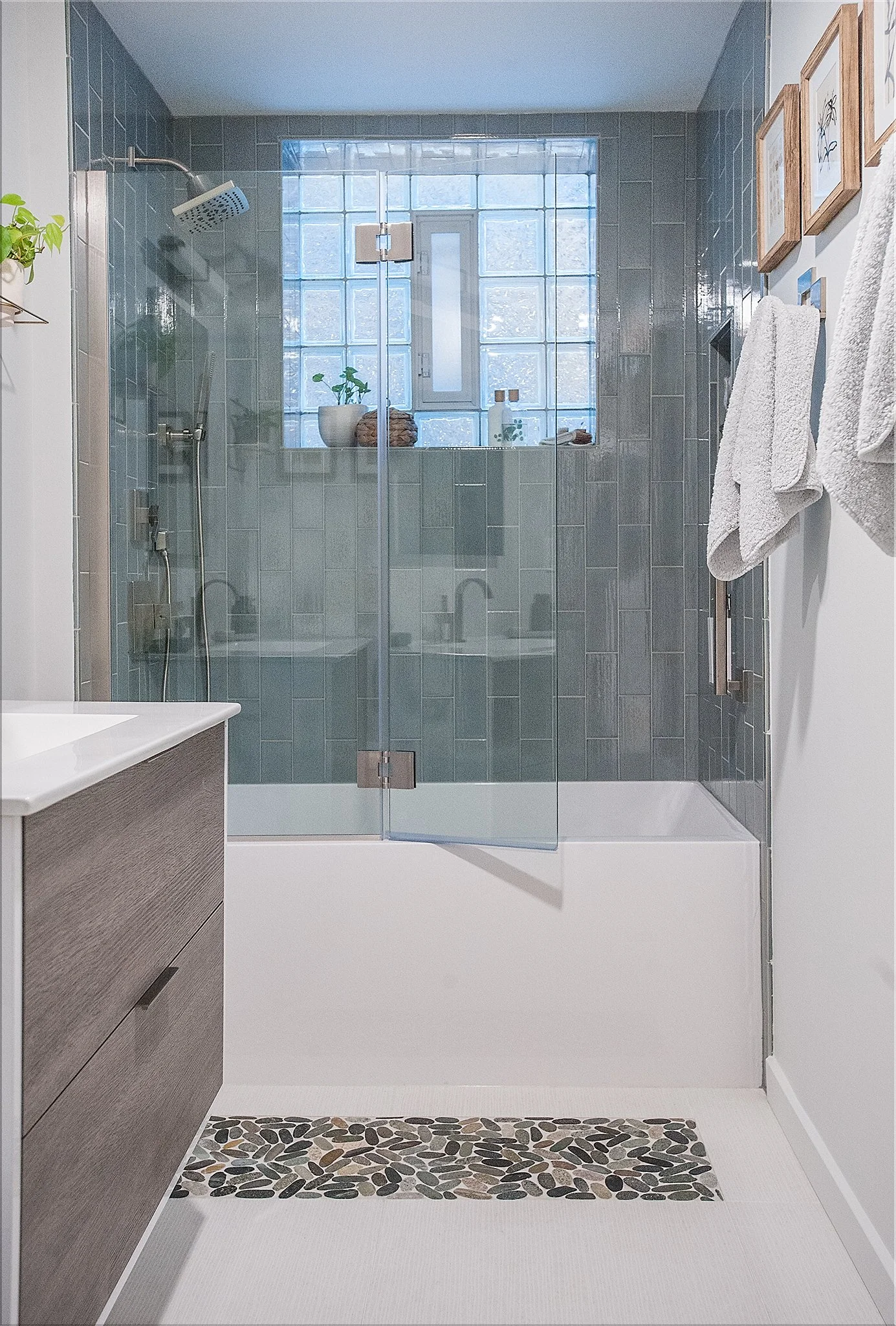 Modern bathroom with a glass shower enclosure, dark gray tiles, a window with glass blocks, and white towels hanging on the wall.