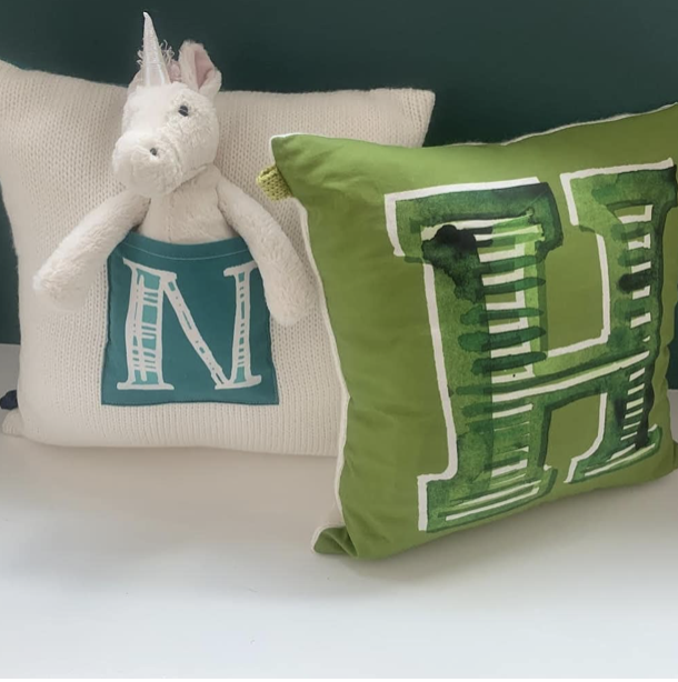 A plush unicorn toy with a party hat sitting on a cream pillow with a large letter 'N' on it, next to a green pillow with a large letter 'H' on it.