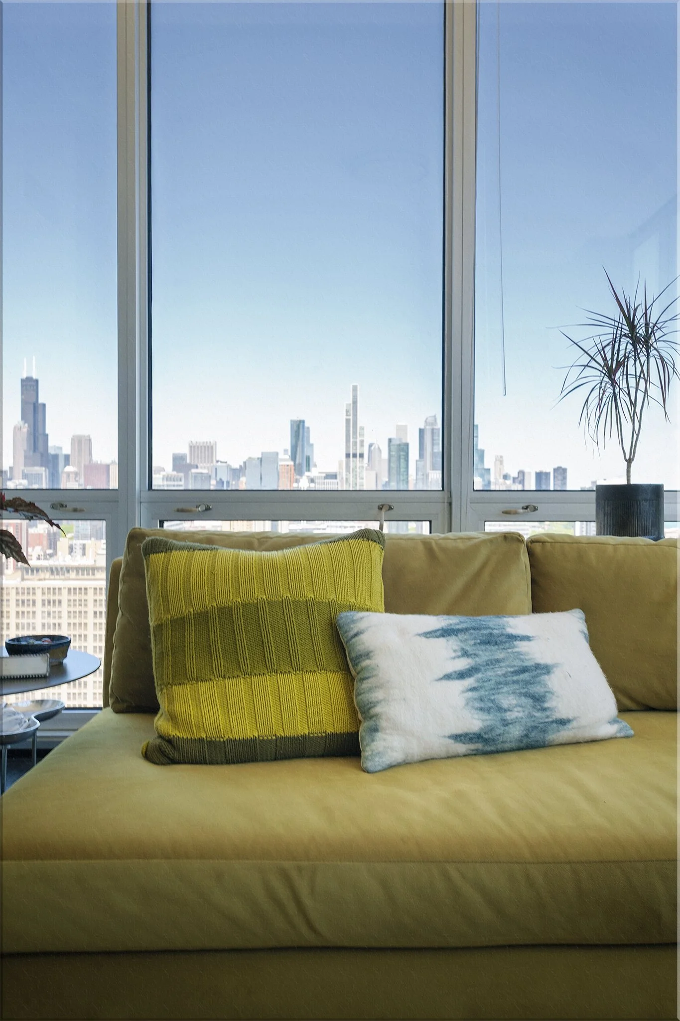 Interior view of a living room with a yellow sofa, decorative pillows, and large windows showing a city skyline.