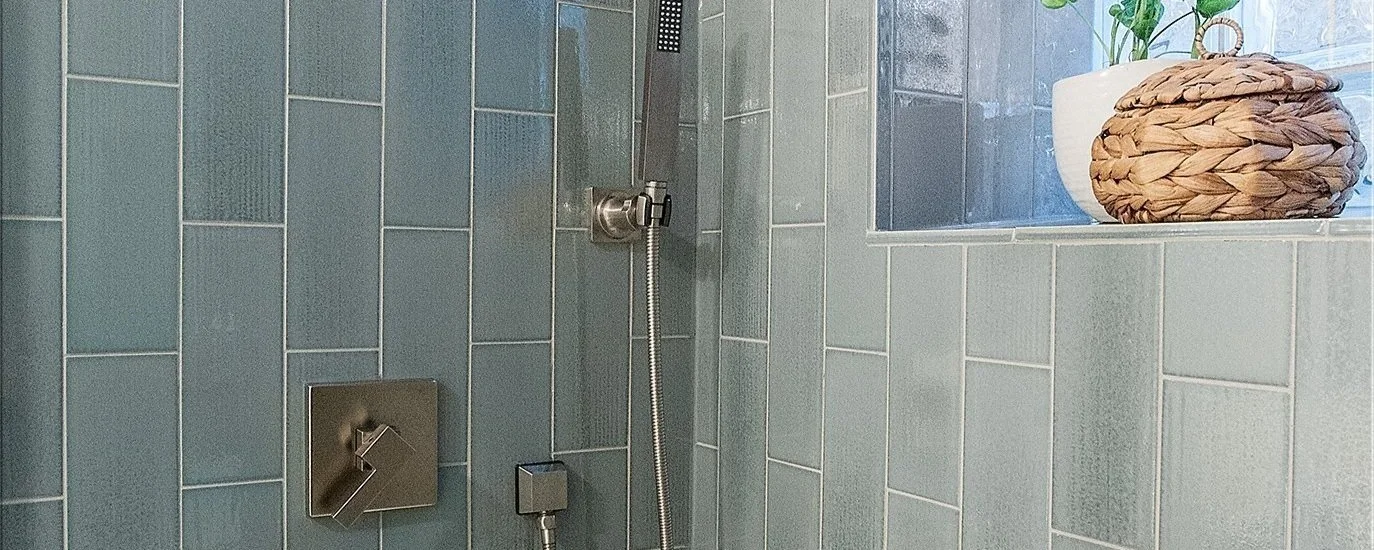 Modern shower with blue tiles, a rain showerhead, a handheld shower wand, a small shelf with a plant and a woven basket next to a frosted glass block window.