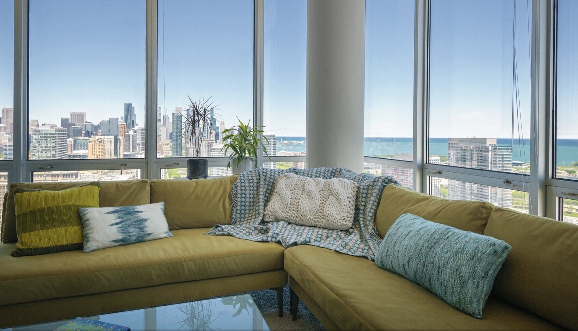 Living room with mustardsectional sofa, threw blankets, and city skyline view through large windows, overlooking Chicago lakeshore.