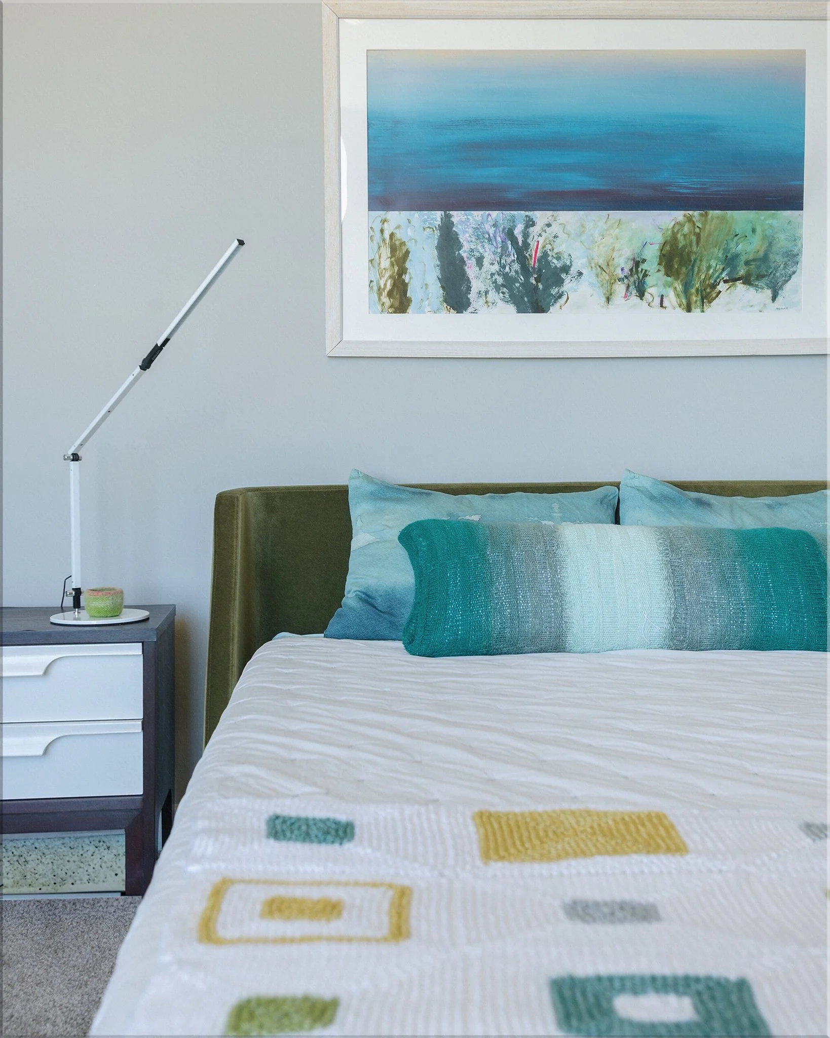 A bedroom with a bed featuring teal pillows and a quilt with yellow and teal geometric patterns. A white nightstand with a circular lamp and a small object sits next to the bed. A framed abstract landscape painting depicting a blue sky, water, and trees hangs on the wall above the bed.