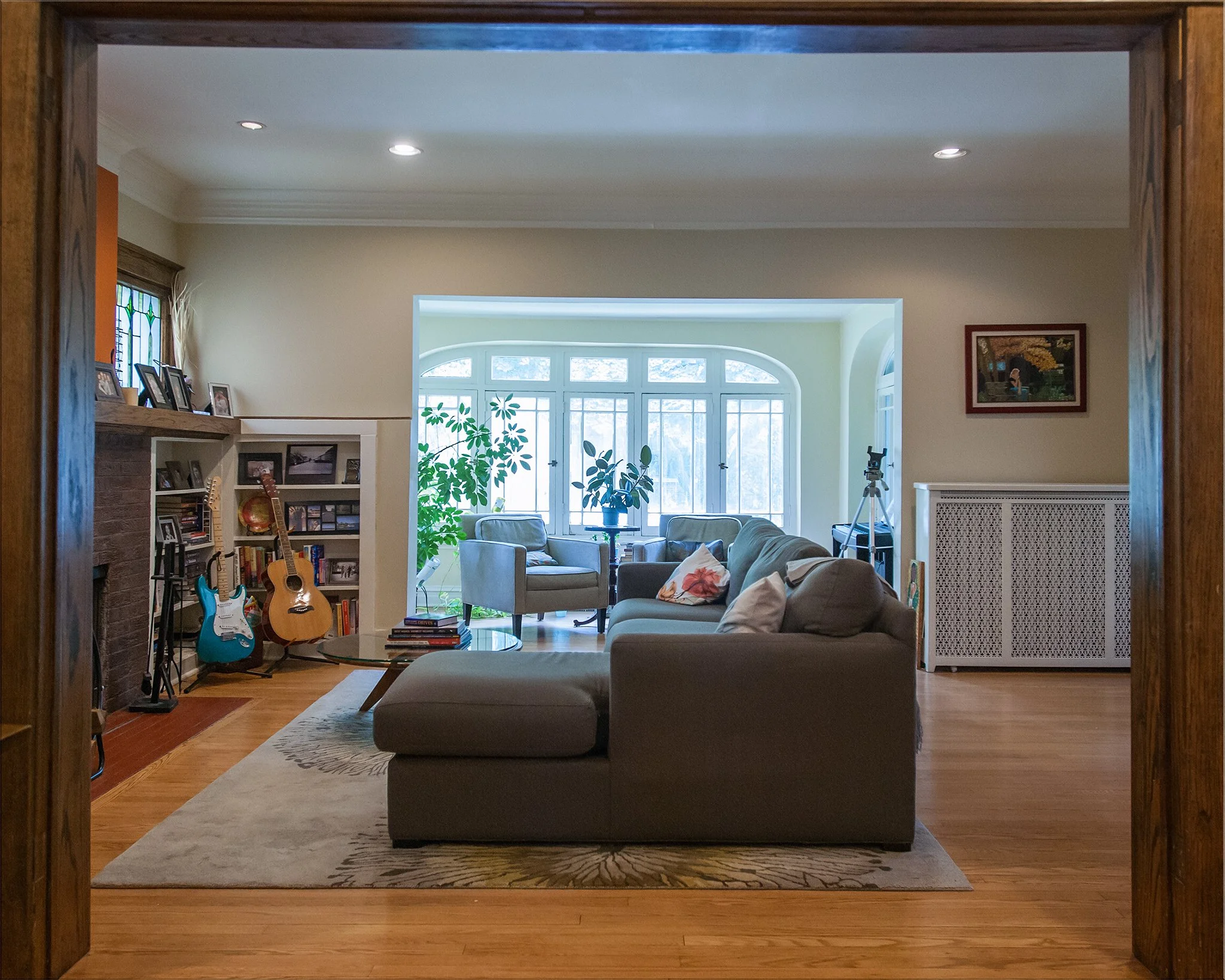 Living room with large window, gray sofa, armchairs, bookshelf with guitars, and indoor plants.