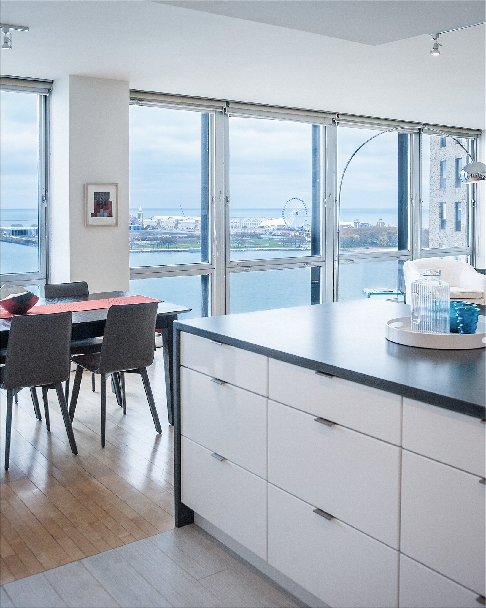 Modern apartment interior with a dining table and chairs, large windows showing a water view, and a kitchen island with decorative items.