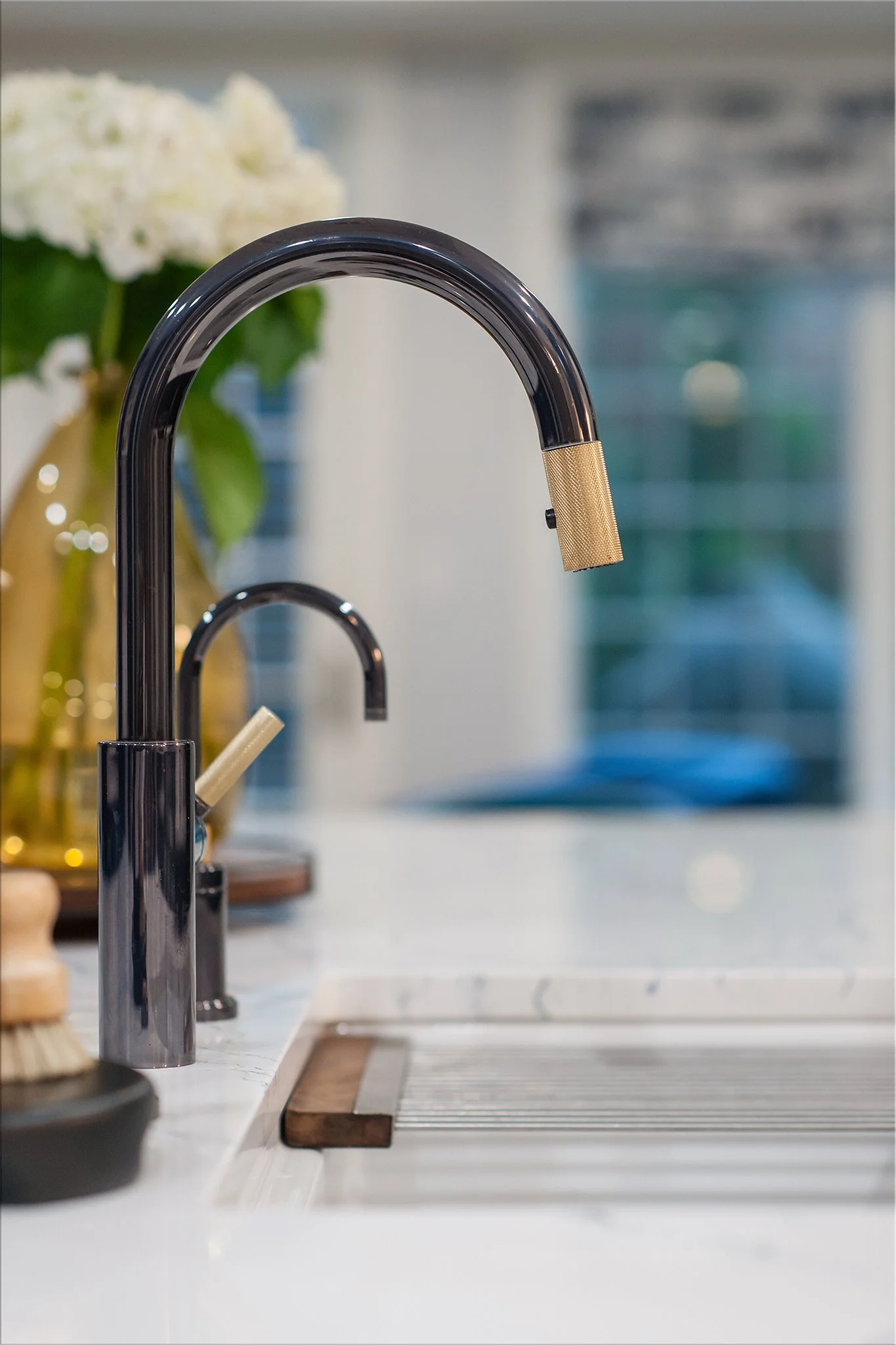 Close-up of a modern black kitchen faucet with a gold handle and spray head, on a white marble countertop, with a flower arrangement and a window in the background.