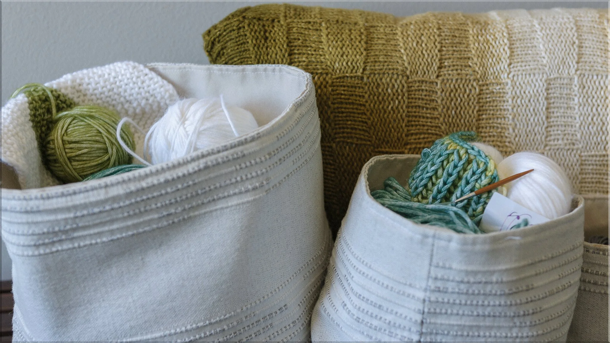 Two fabric storage baskets filled with balls of yarn, knitting needles, and a white knitted item, placed in front of a yellow and beige woven cushion.