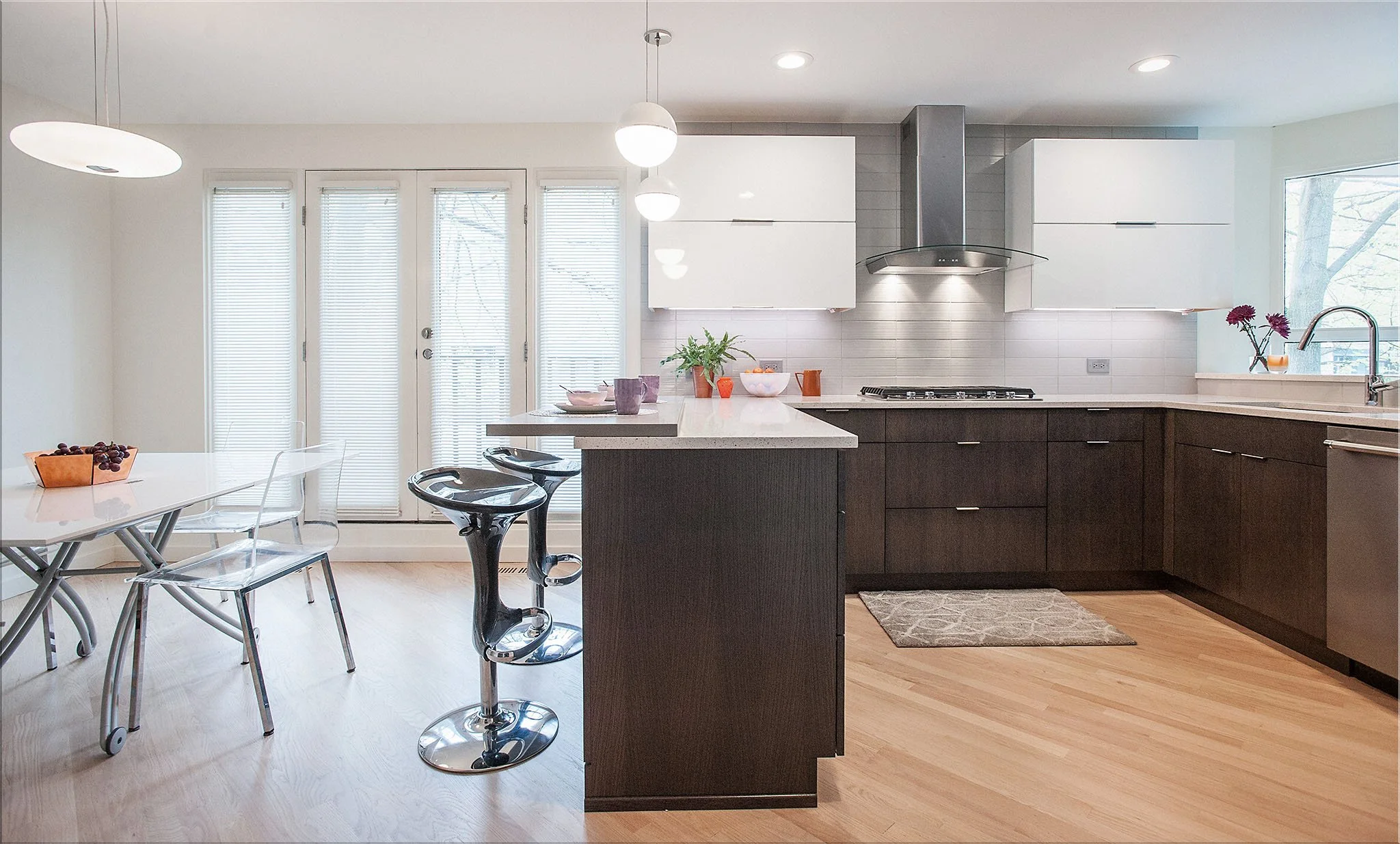 Modern kitchen with white upper cabinets, dark lower cabinets, a central island with a white countertop, stainless steel appliances, a round dining table with transparent chairs, bar stools, large windows, and hardwood floors.