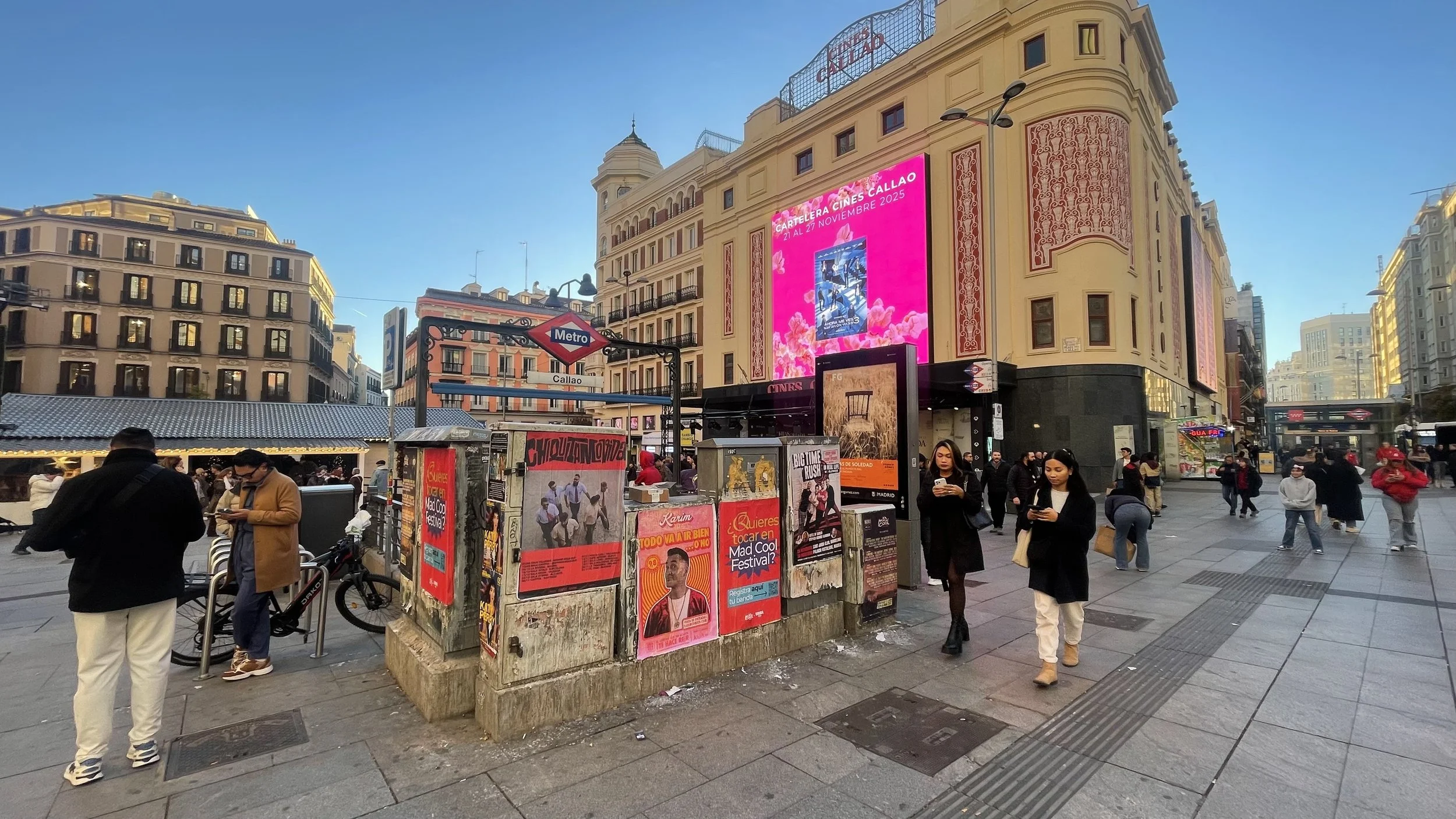 Busy city street with people walking, a large digital billboard, posters on a wall, and a Metro sign. Buildings surround the scene with a clear sky overhead.