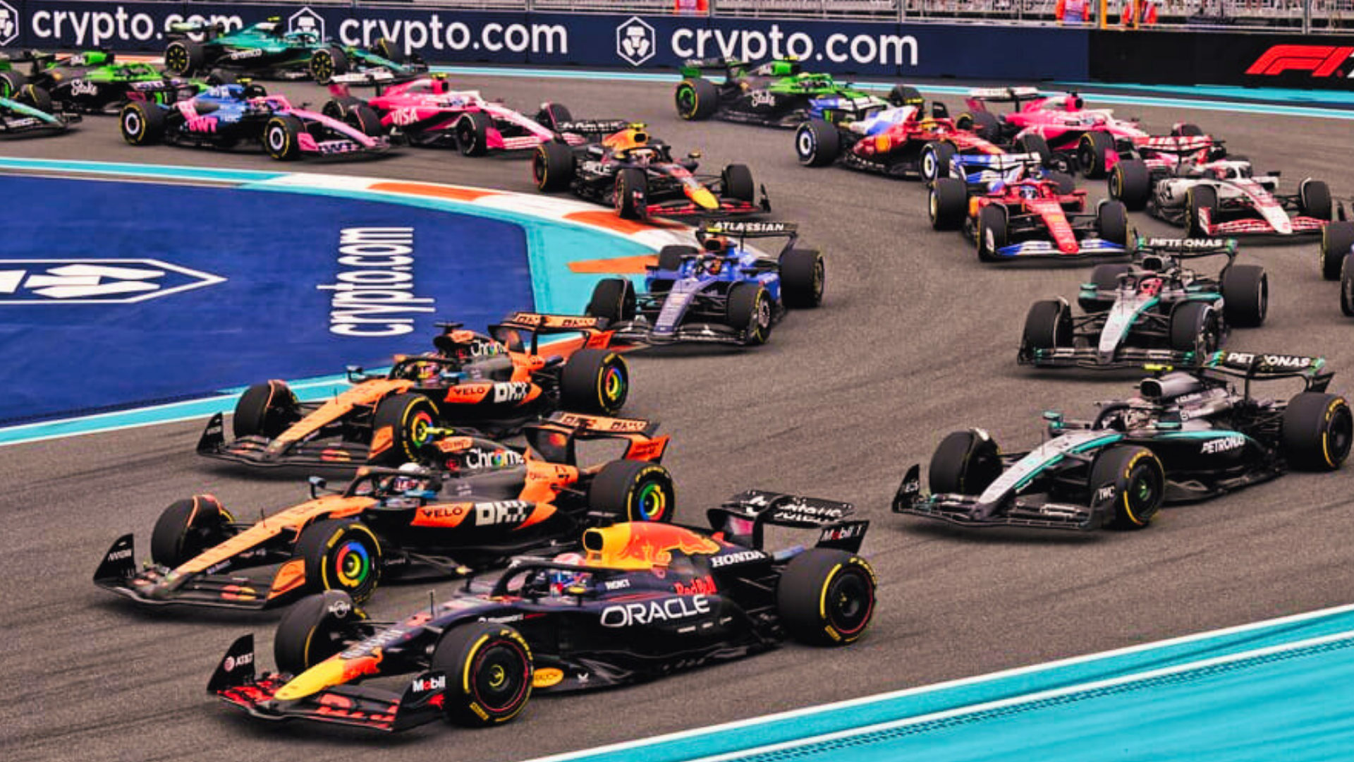 Multiple Formula 1 race cars on a track during a race with branding visible in the background.