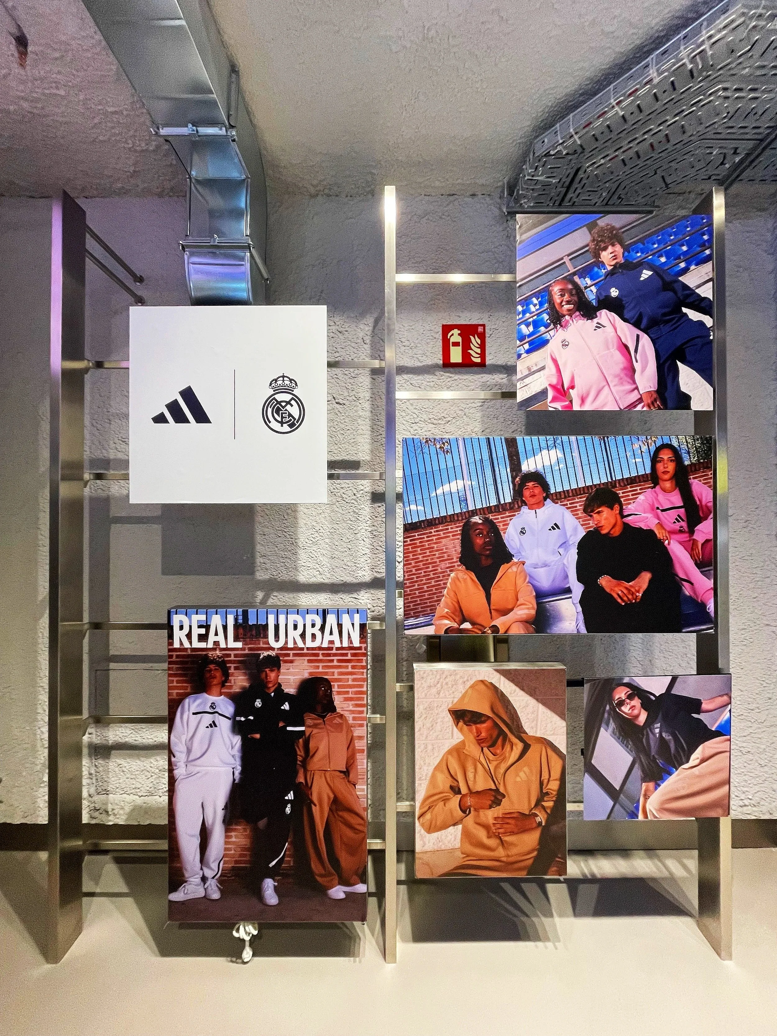 Display featuring photos of young diverse individuals in casual and sportswear, with branding for Adidas and Real Madrid, on a metal frame with a fire extinguisher sign in the background.