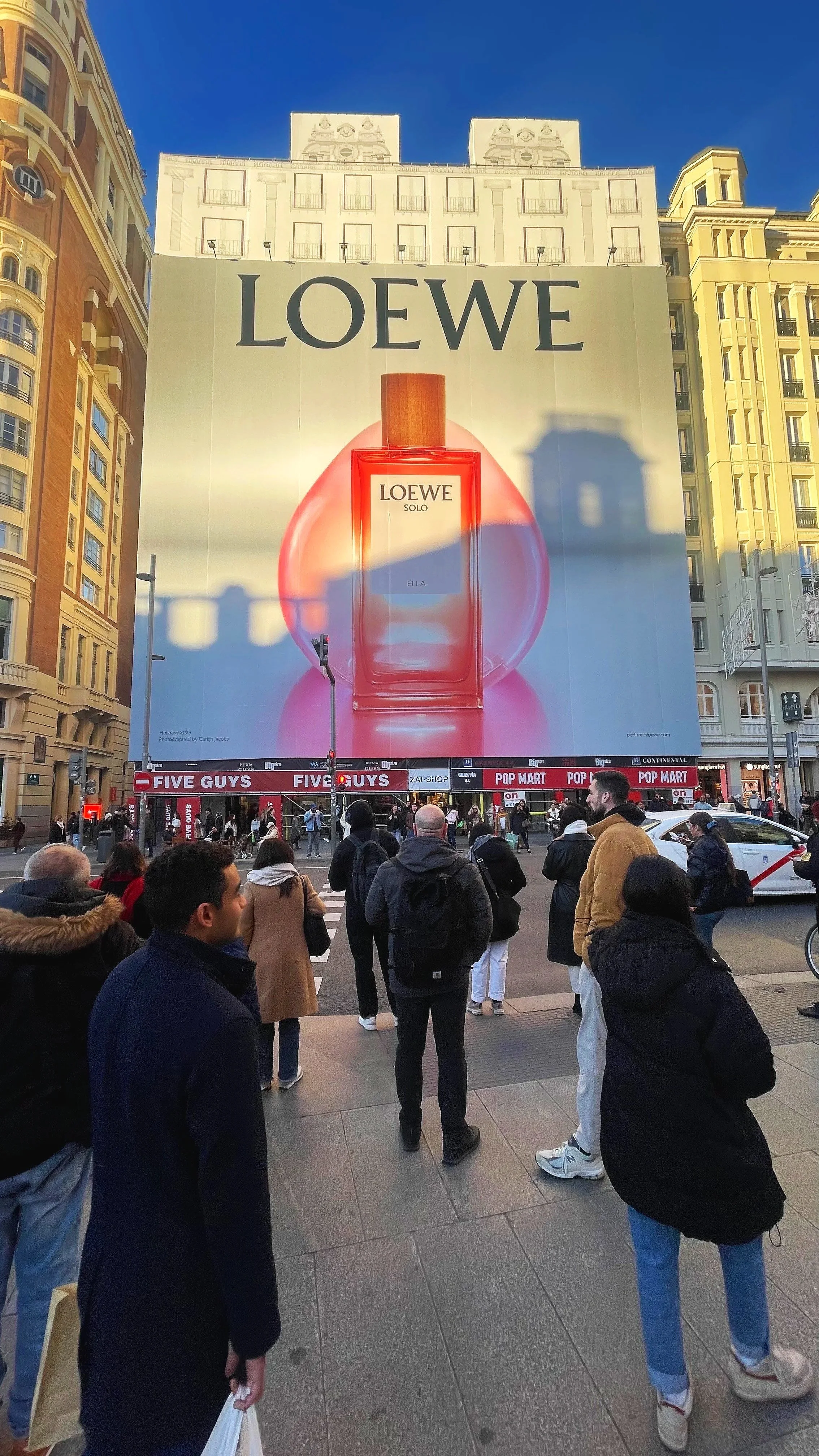 Large billboard advertising Loewe Solo Ella perfume with a red perfume bottle image at the center. The billboard is on a city street with people waiting at the crosswalk, and surrounding buildings.
