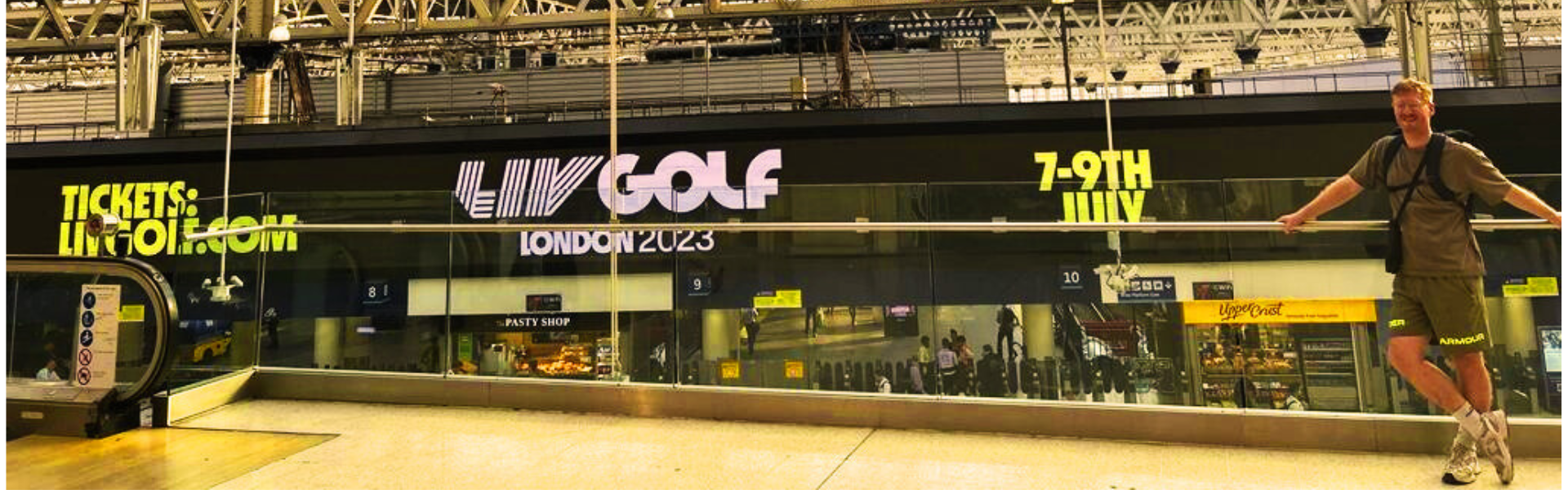 A man standing and smiling at an airport terminal with a large sign advertising LIV Golf in London, July 7-9, 2023, with a pastry shop visible in the background.