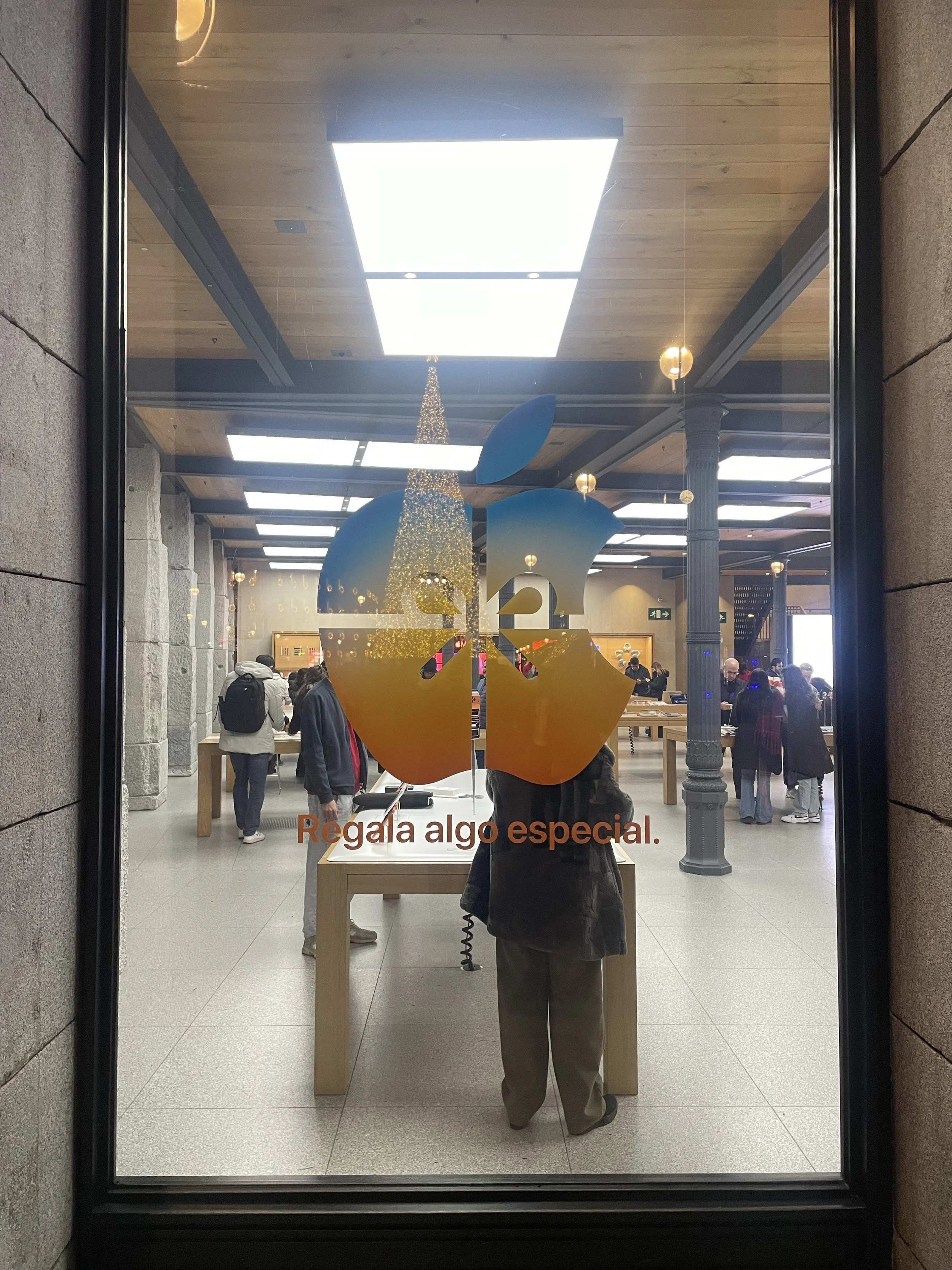 Inside a store viewed through a glass window with a large apple logo and the Spanish phrase "Regala algo especial." The store has people shopping, wooden tables, stone walls, and lighting fixtures.