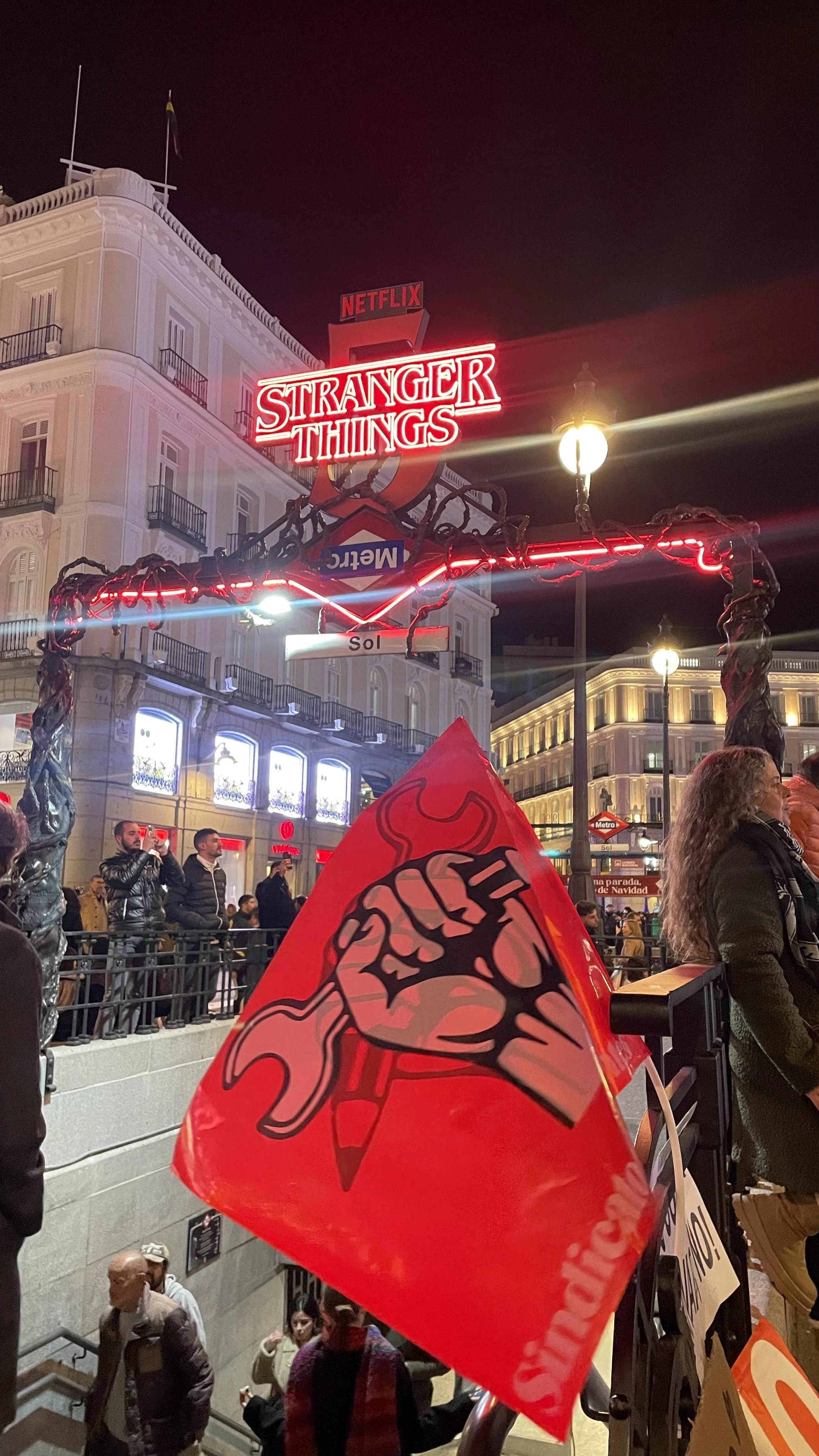 Nighttime scene in a city with a crowd of people. Prominent neon sign reads 'STRANGER THINGS' with Netflix logo above. Red flag with a raised fist holding a pen, symbolizing activism, is in the foreground.