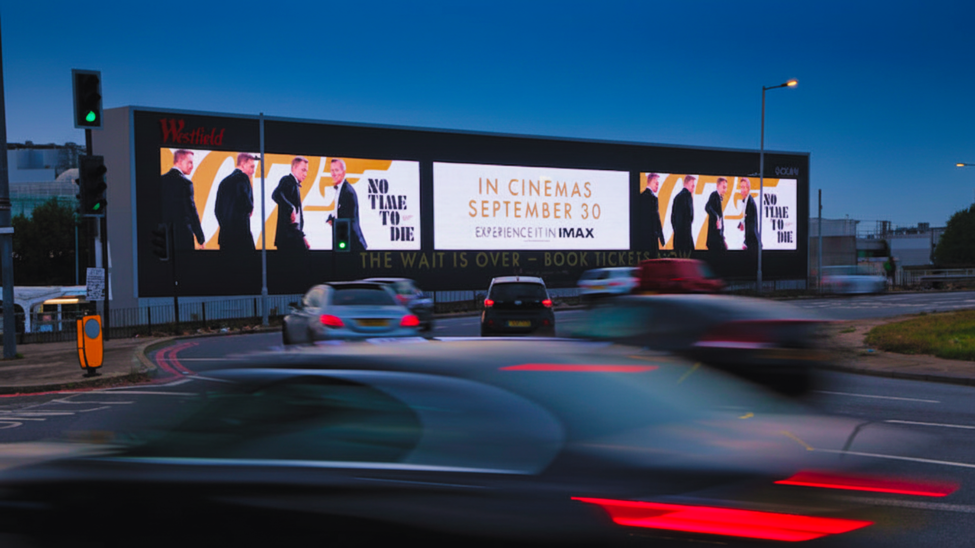 A digital billboard advertising the movie 'No Time to Die,' with release date September 30, and the message 'Experience it in IMAX,' located on a busy street at dusk, with cars passing by.