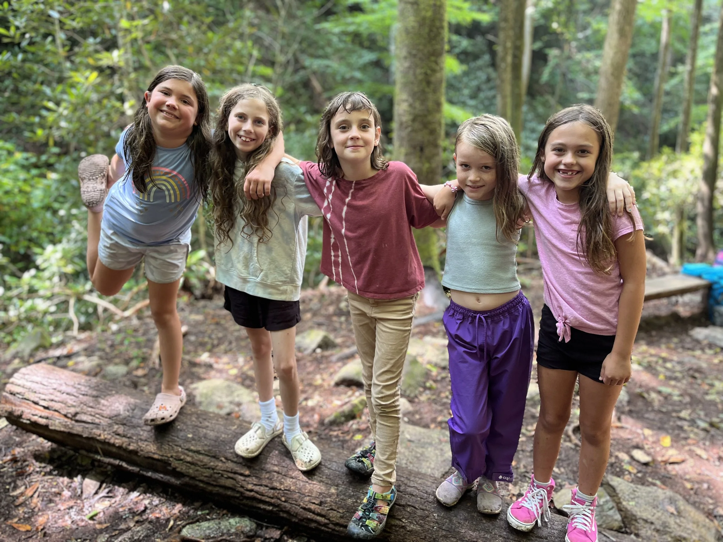 Six young girls standing on a fallen log in a forest, smiling at the camera with their arms around each other.