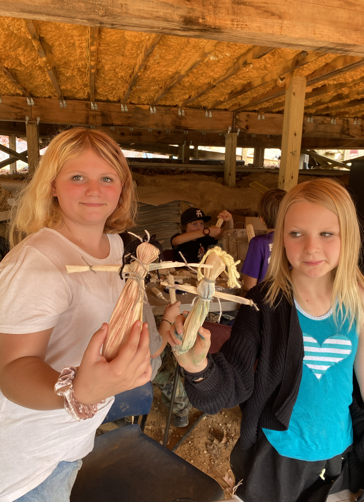 Two young girls displaying corn husk dolls they made, standing under a wooden shelter with other children in the background.