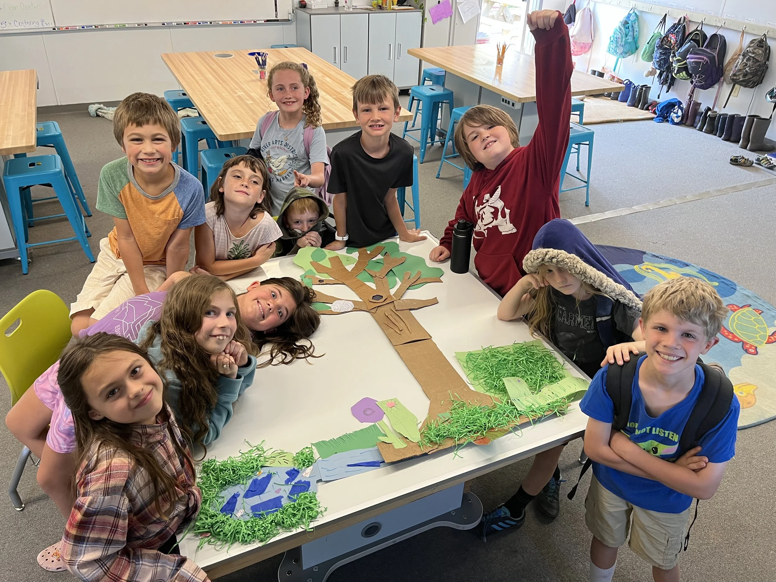 Group of children gathered around a table displaying a large paper tree craft with green paper grass and trees, inside a classroom.