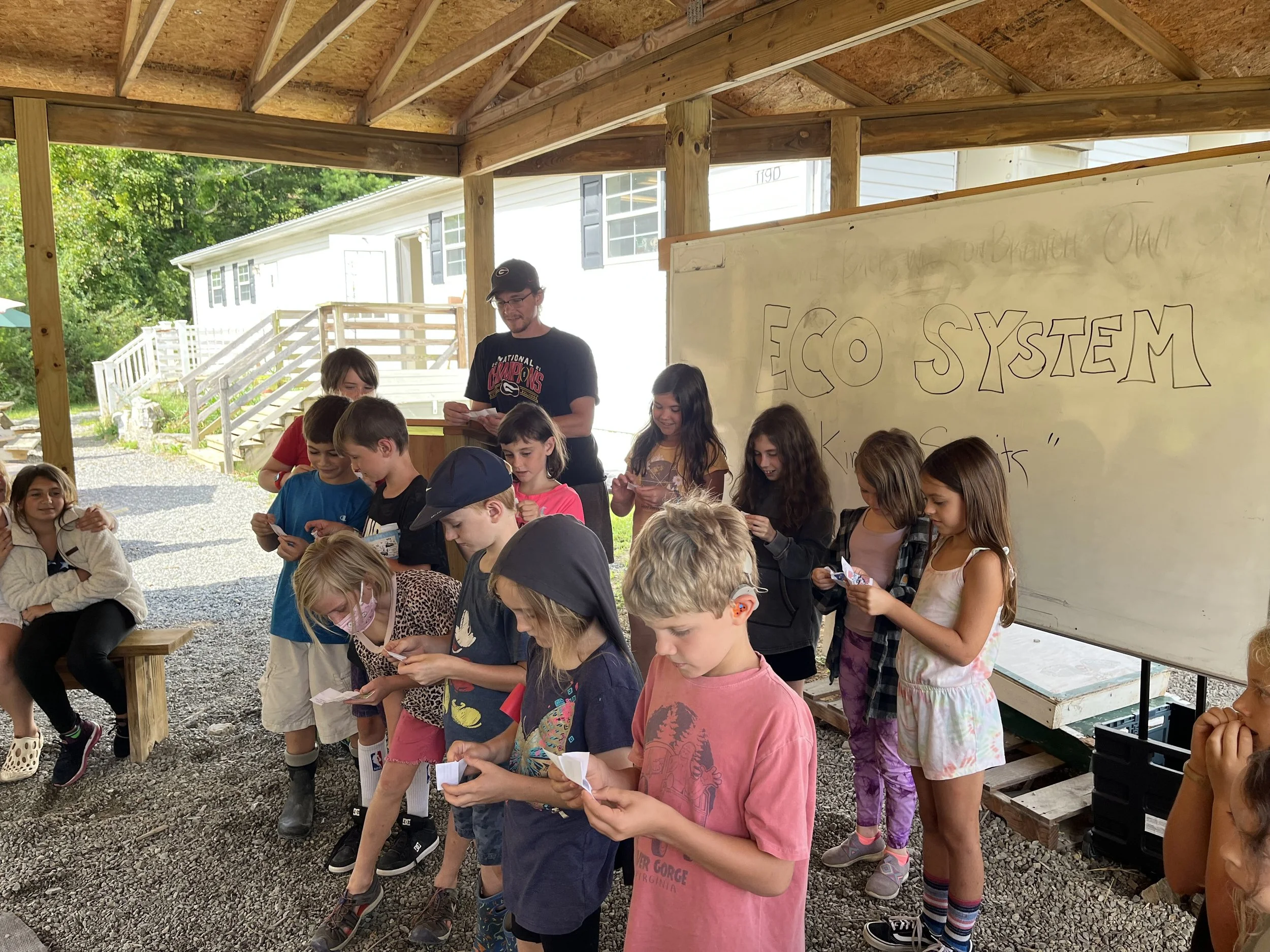 Children gathered under a wooden shelter, looking at notes or papers, with a whiteboard in the background reading 'ECO SYSTEM' and other writing at a summer camp or outdoor classroom.