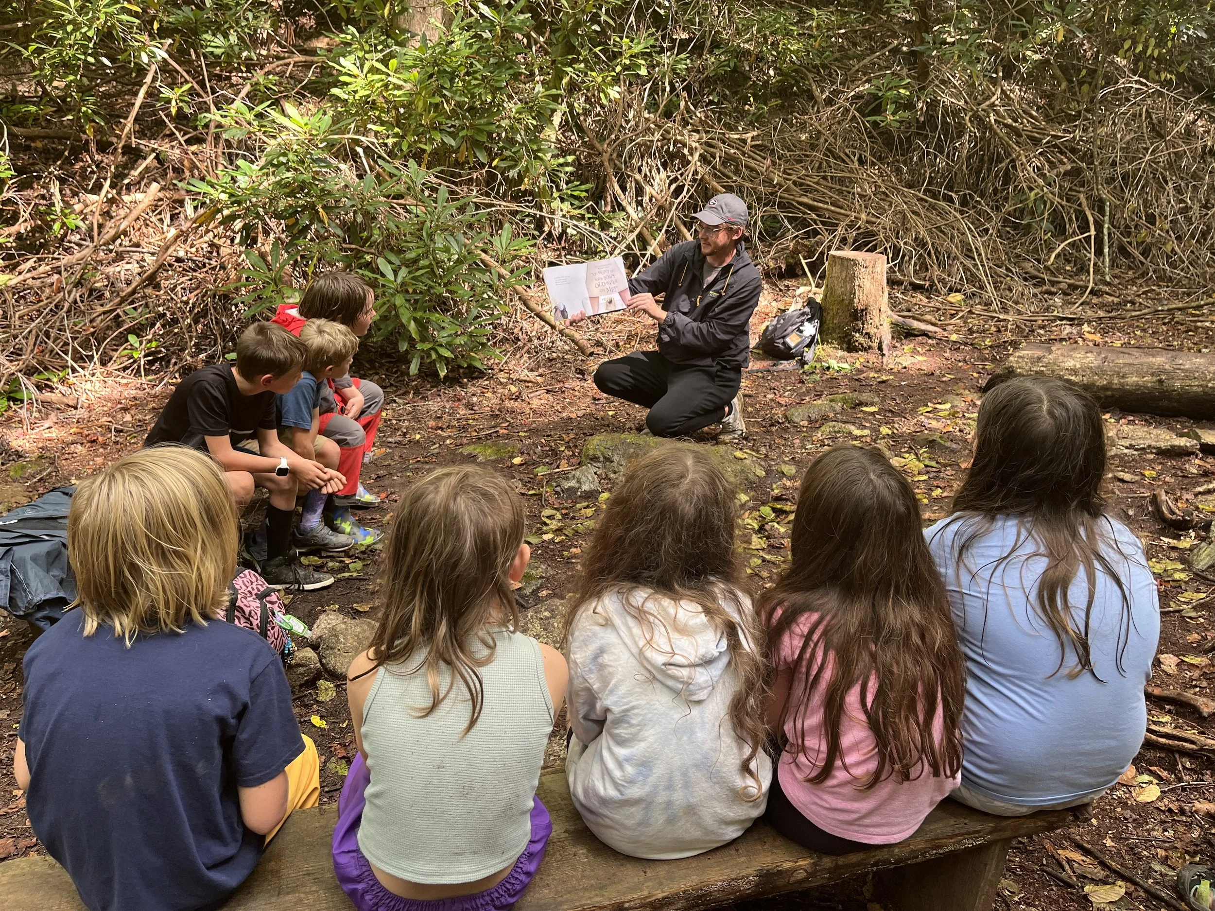 A man kneeling and reading a book aloud to a group of children sitting on a log in the woods.