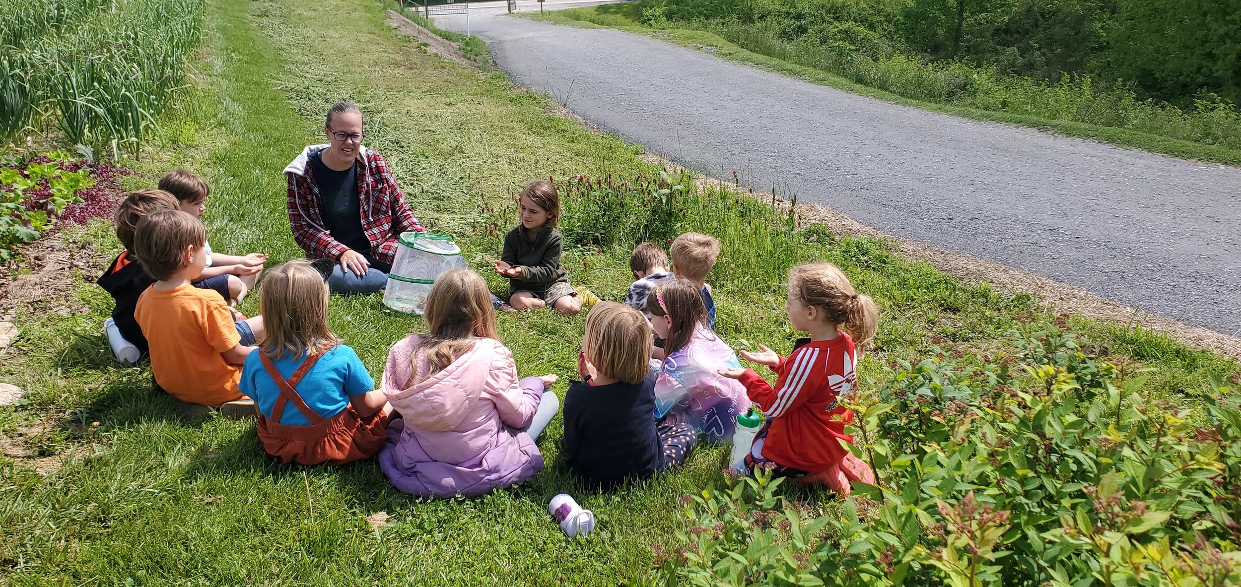 A group of children sitting on the grass near the roadside, listening to a woman who appears to be teaching or talking to them, on a sunny day.