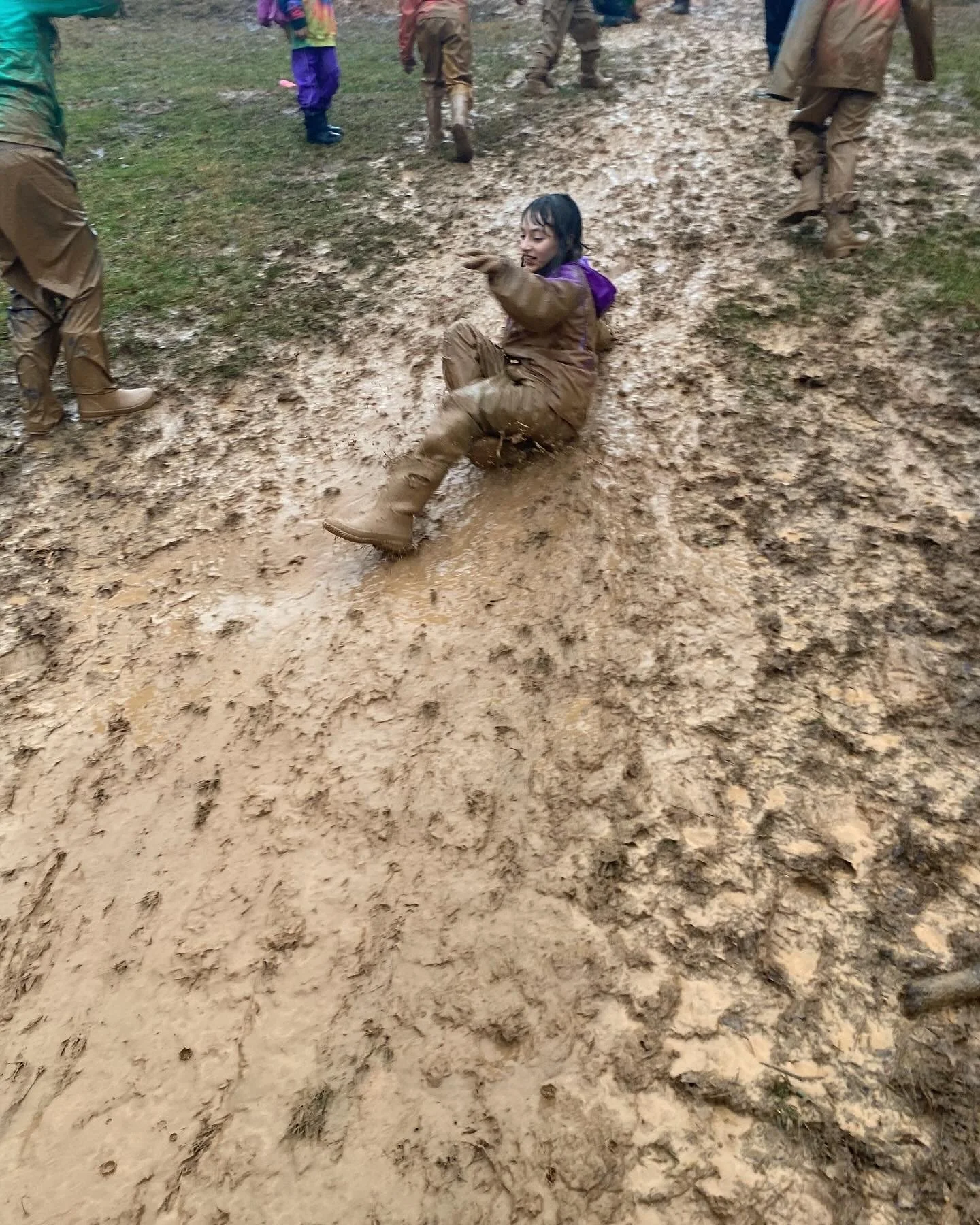 A young girl in muddy clothing sitting on a muddy slope in a muddy outdoor area, surrounded by other people also dressed in muddy clothing, possibly after sliding or falling on the wet ground.