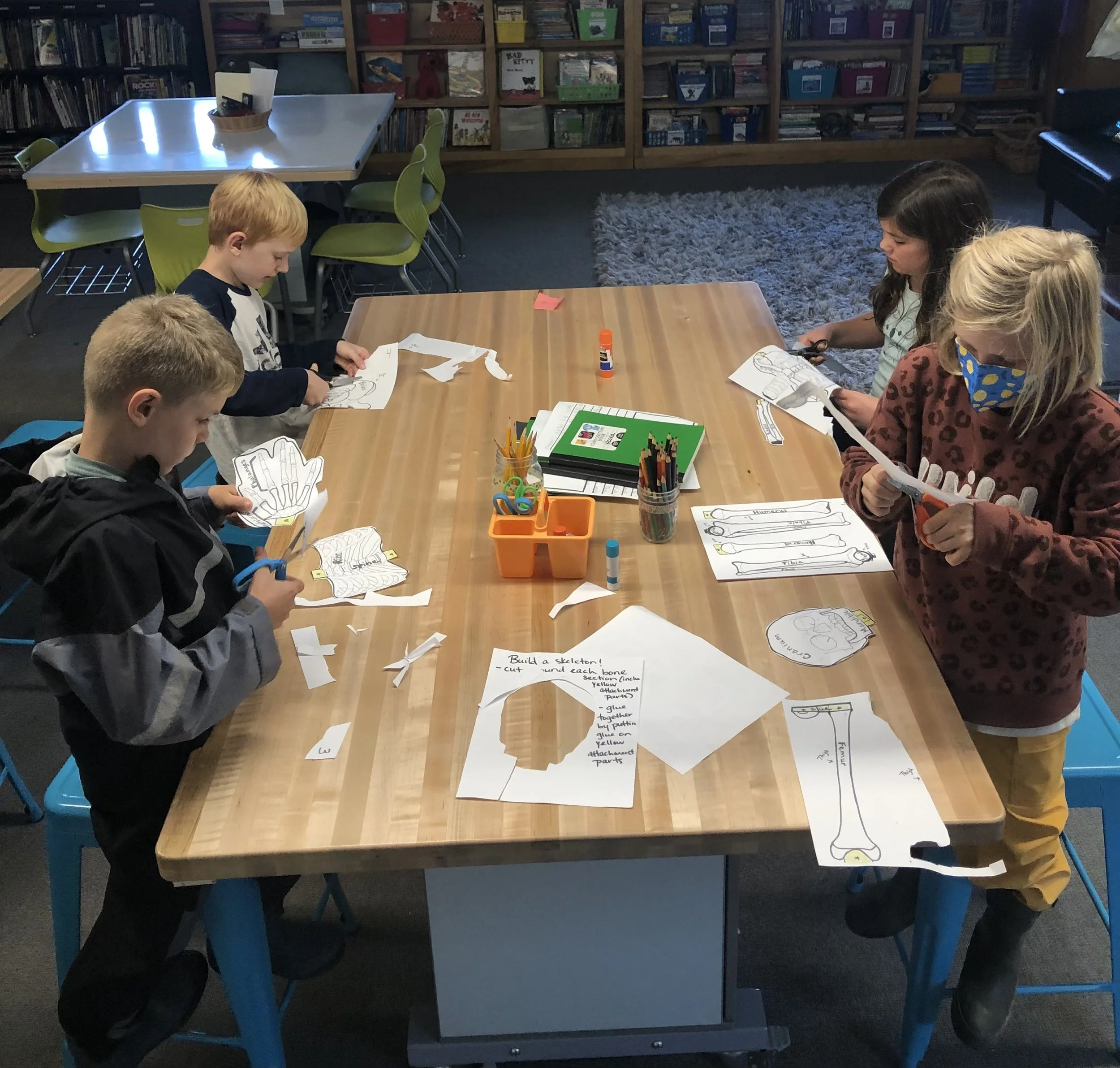 Children engaged in a paper craft activity at a wooden table in a classroom or library, cutting out paper skeletons with scissors and gluing parts together, surrounded by art supplies.
