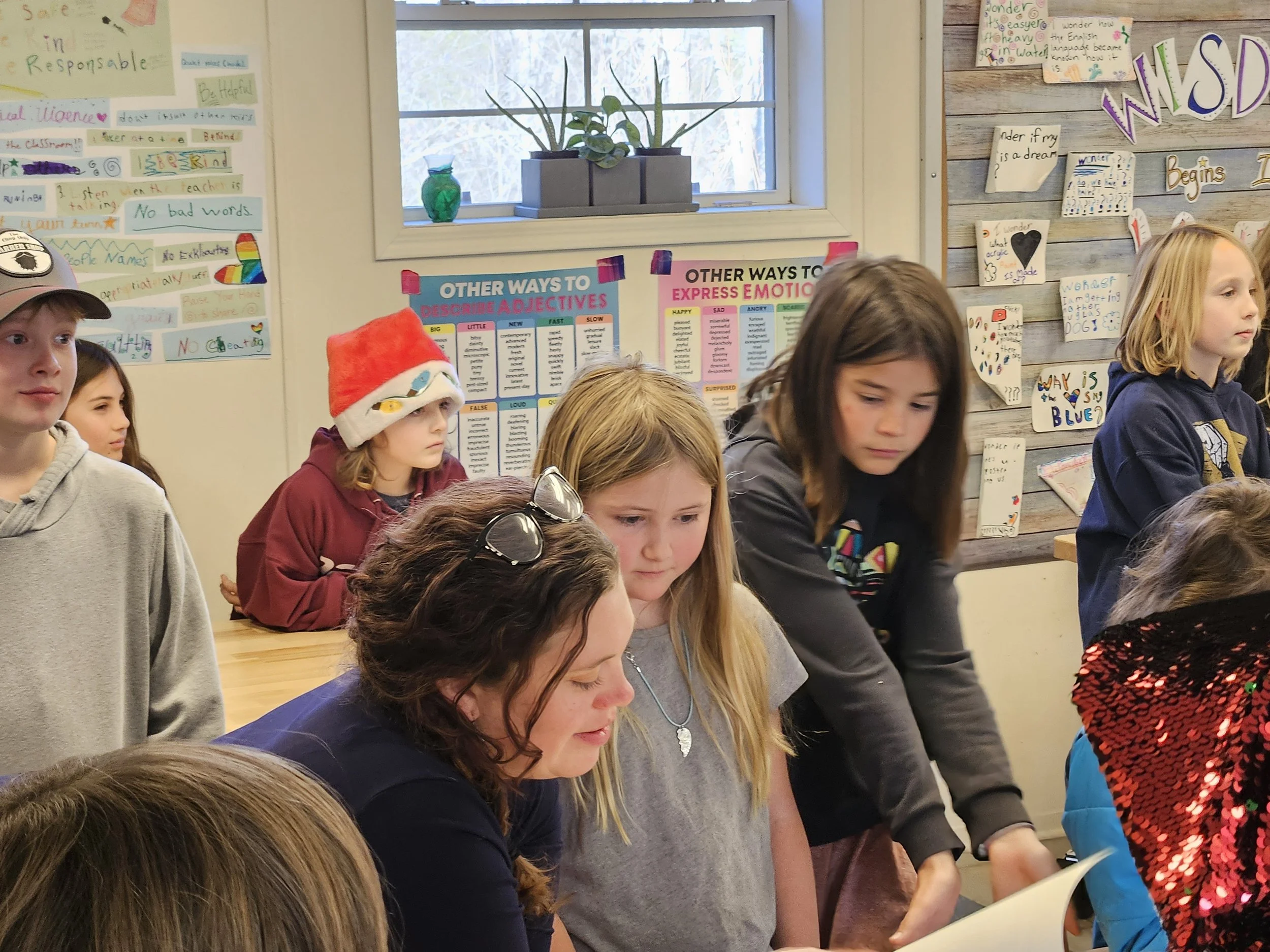 A group of children and a woman gathered around a table in a classroom, with posters and artwork on the walls.