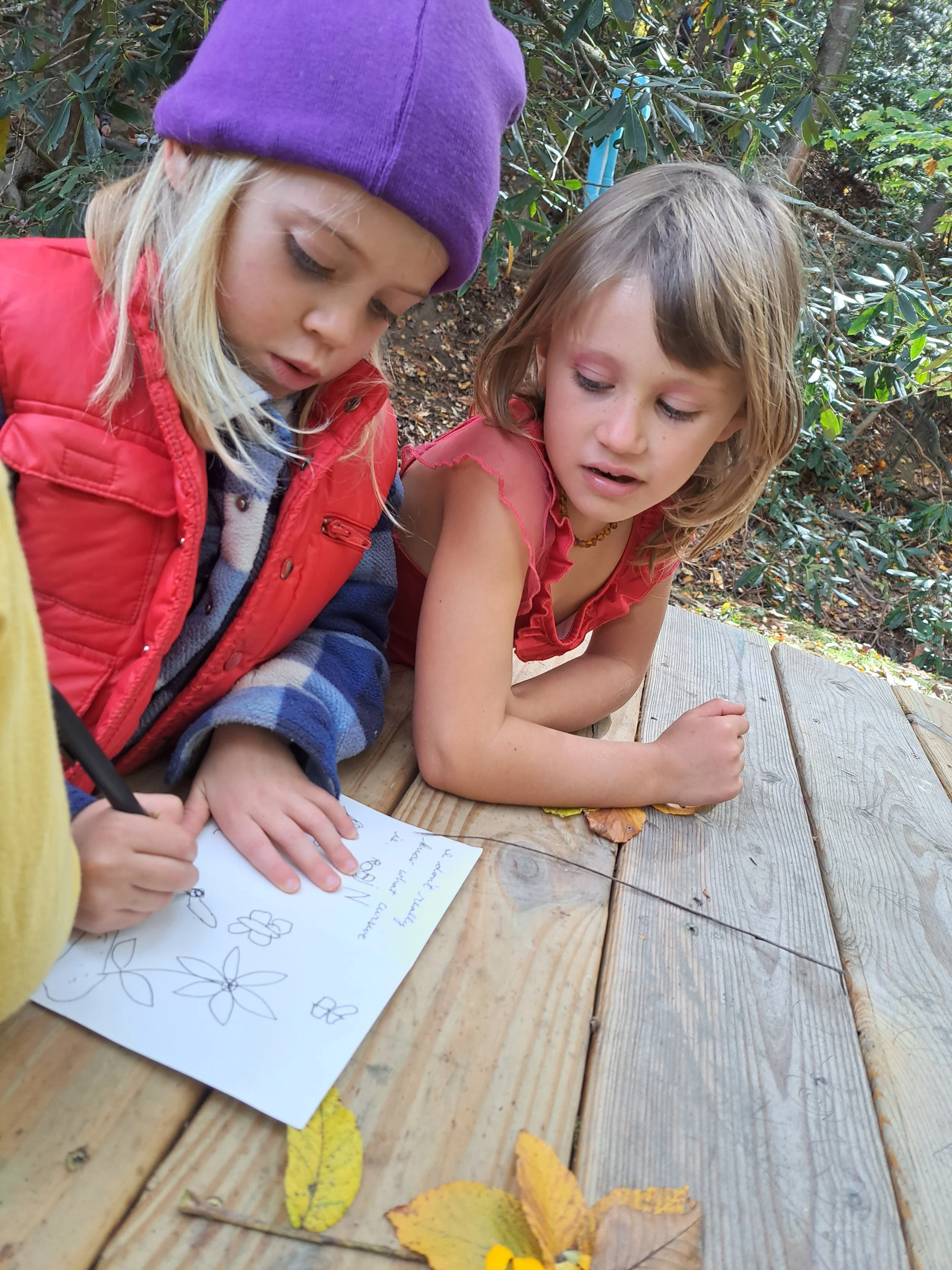 Two young girls looking at a piece of paper with drawings of flowers, sitting at a wooden table outdoors surrounded by green bushy plants and fallen yellow leaves.
