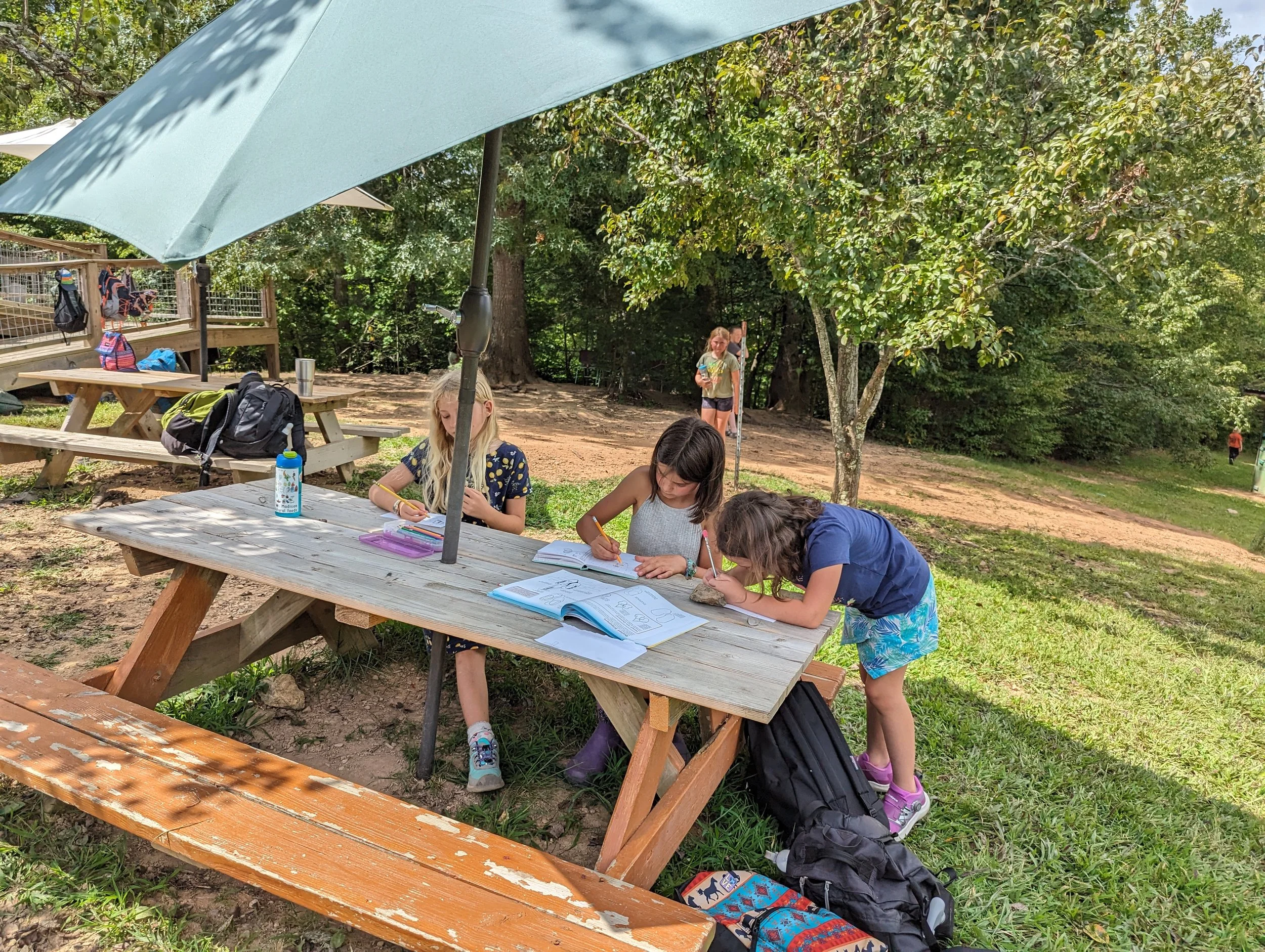 Three children working on homework or drawing at a wooden picnic table outdoors under an umbrella, with backpacks and water bottles nearby, in a park or wooded area.