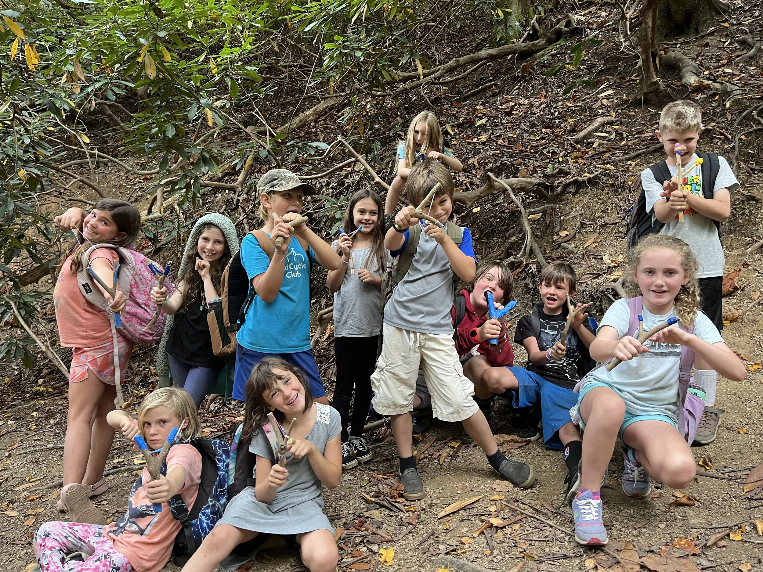 Children on a hiking trip in a wooded area, holding sticks and wearing backpacks, smiling and posing for the camera.