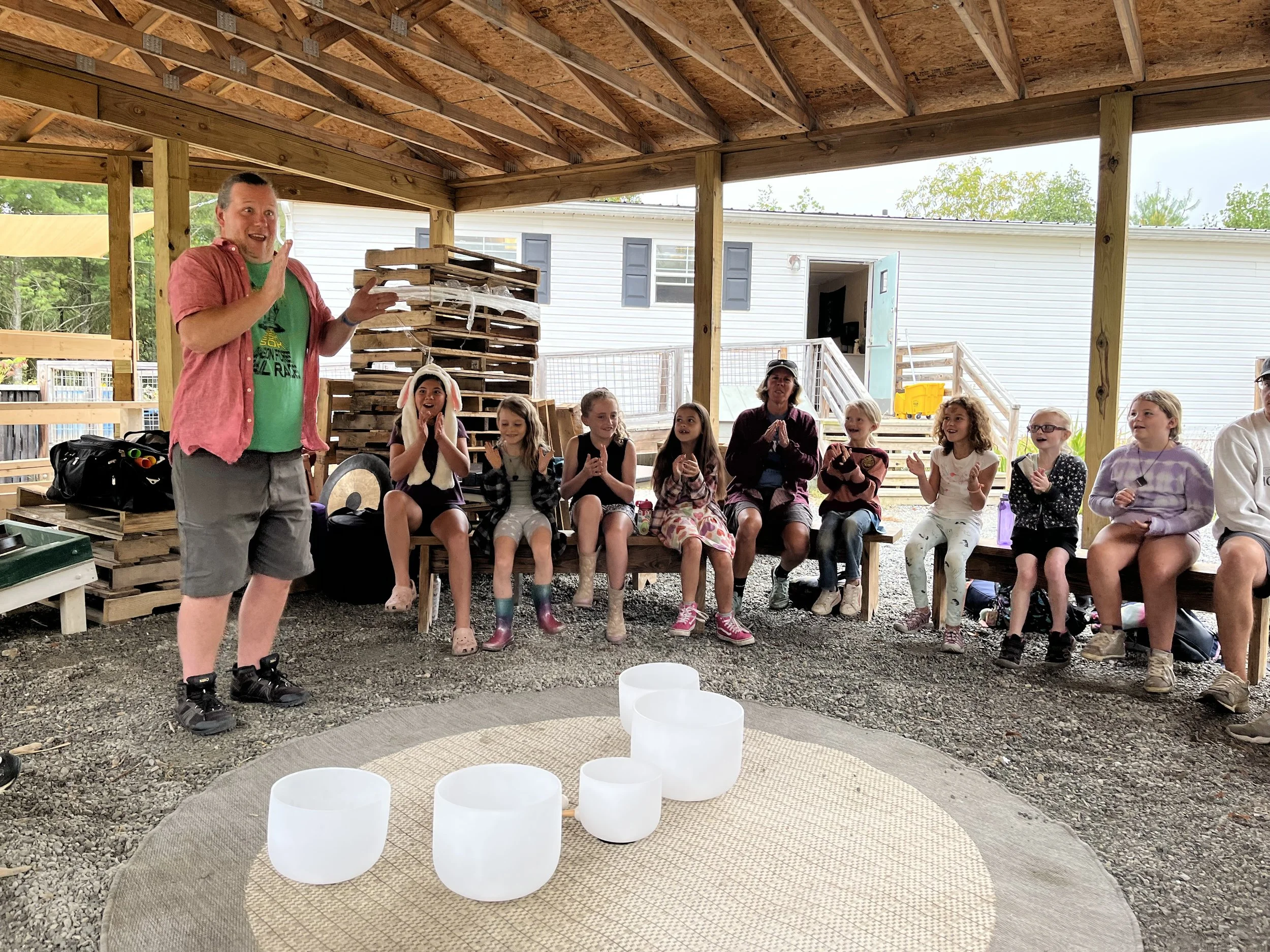A man in a pink shirt and shorts speaks to a group of children sitting on a wooden bench in an outdoor covered area. The children are smiling and clapping, with some wearing rain boots. There are large white singing bowls on a circular mat in the for