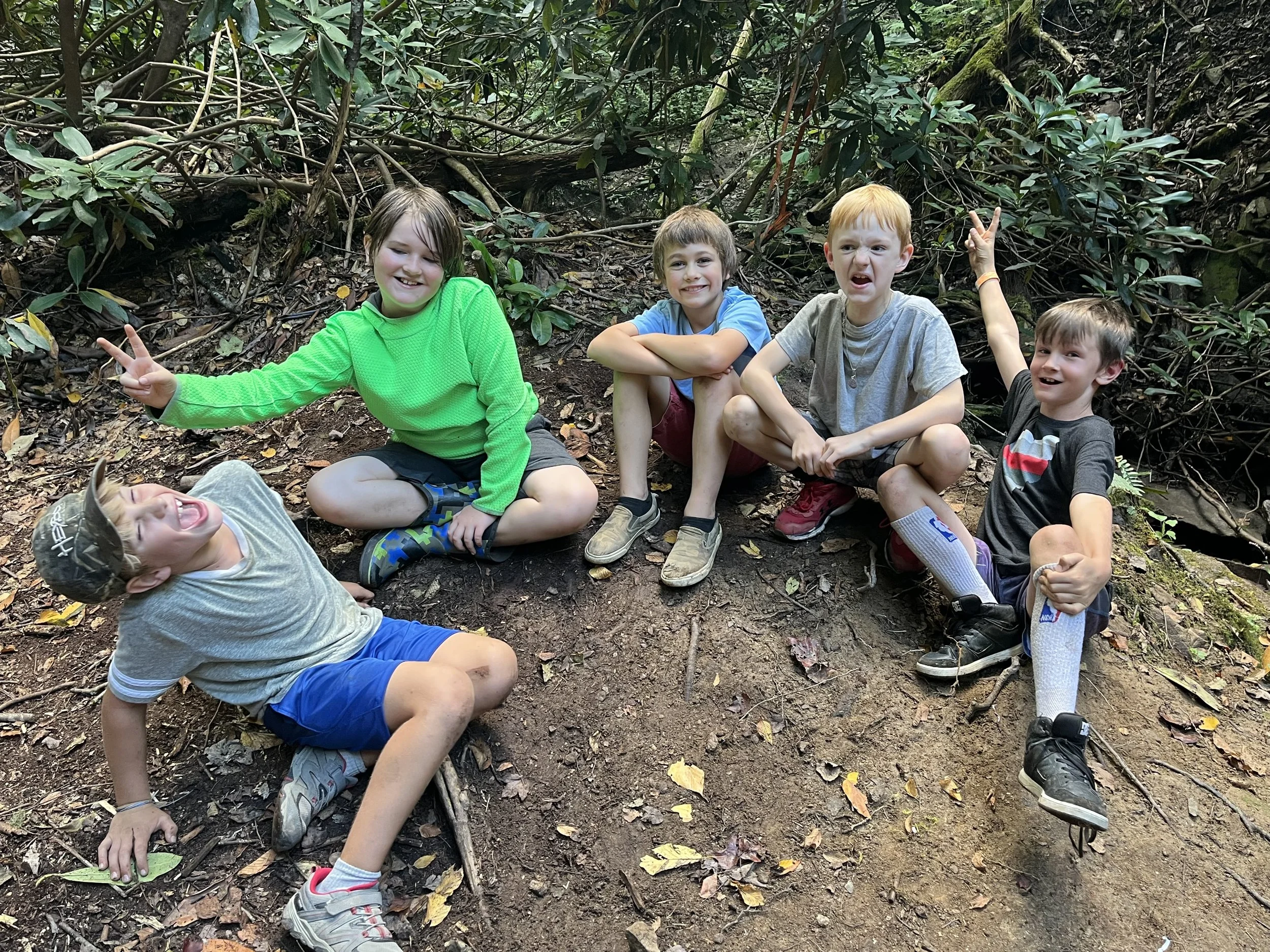 Group of six children sitting and lying on dirt ground in a forest, smiling and making peace signs, surrounded by greenery and fallen leaves.