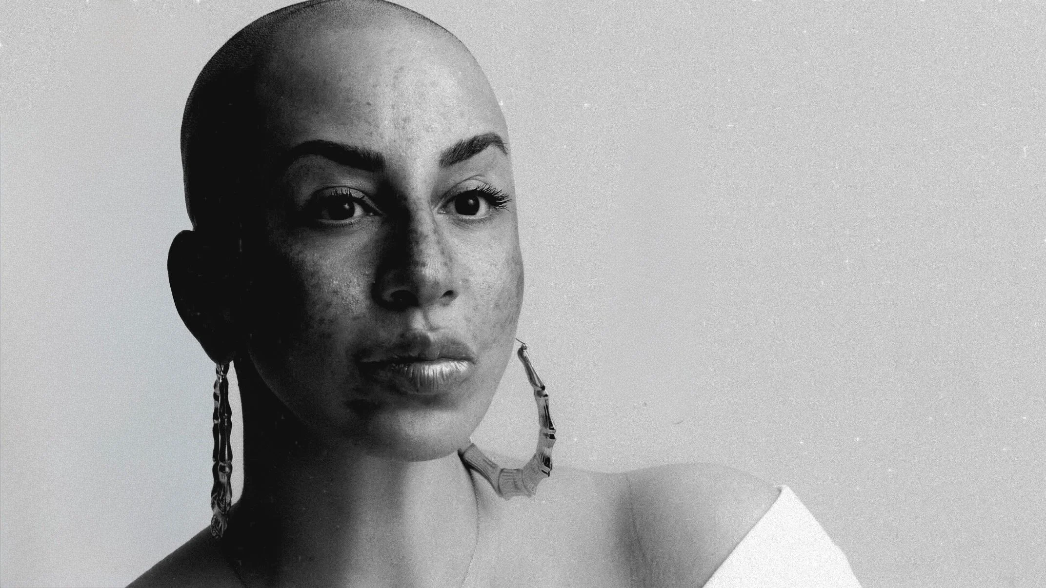 Black and white portrait of a woman with a shaved head, wearing large earrings and a white top, against a plain background.