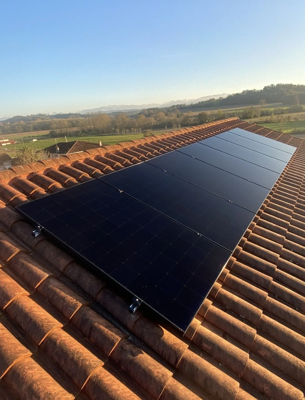 Panneaux solaires installés sur un toit en tuiles, avec un paysage rural en arrière-plan et un ciel clair.
