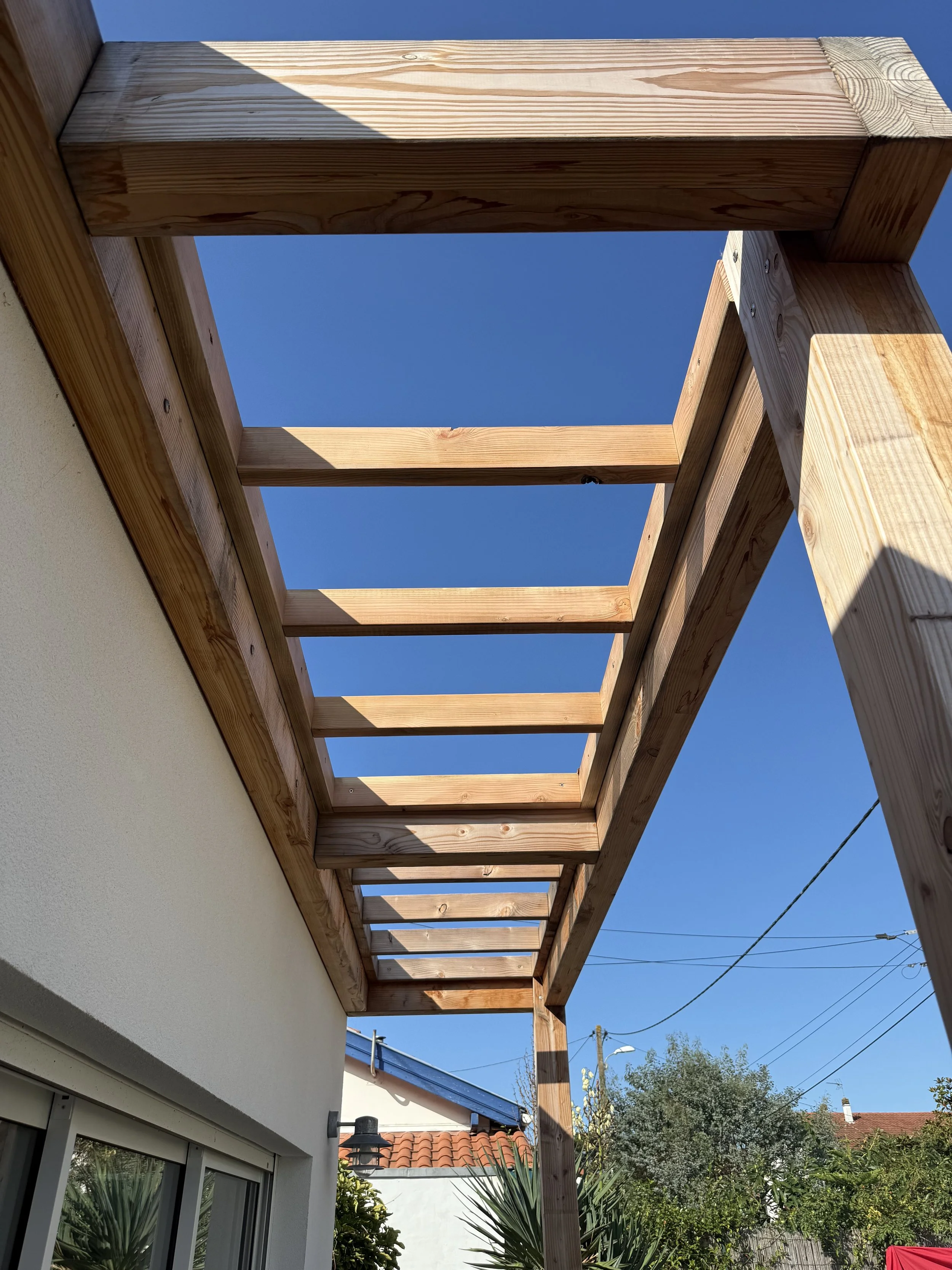 Structure en bois en cours de construction sur la terrasse d'une maison, avec un ciel bleu clair en arrière-plan.