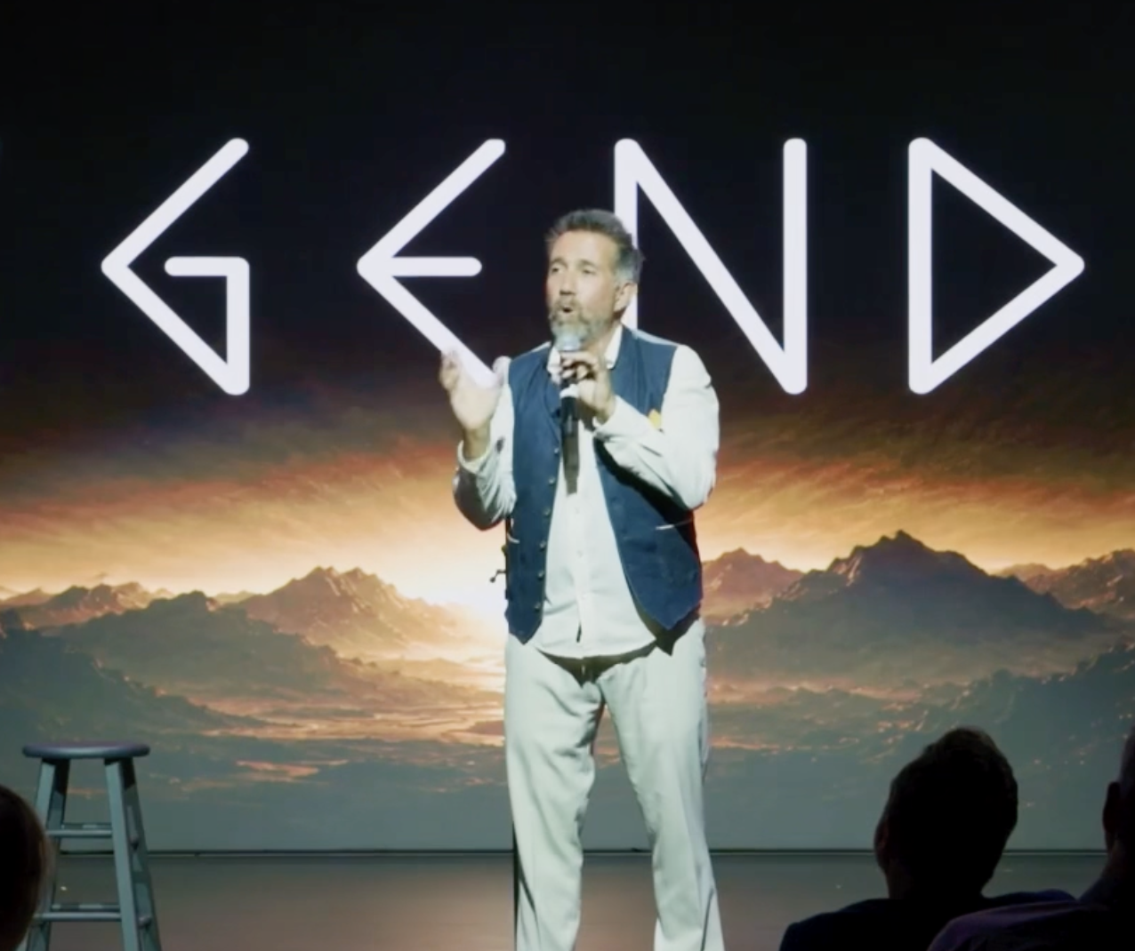 A man standing on stage giving a presentation during a GEND event, with a mountain landscape and sunset scene projected behind him. The word GEND is displayed in large, neon-like letters above him.