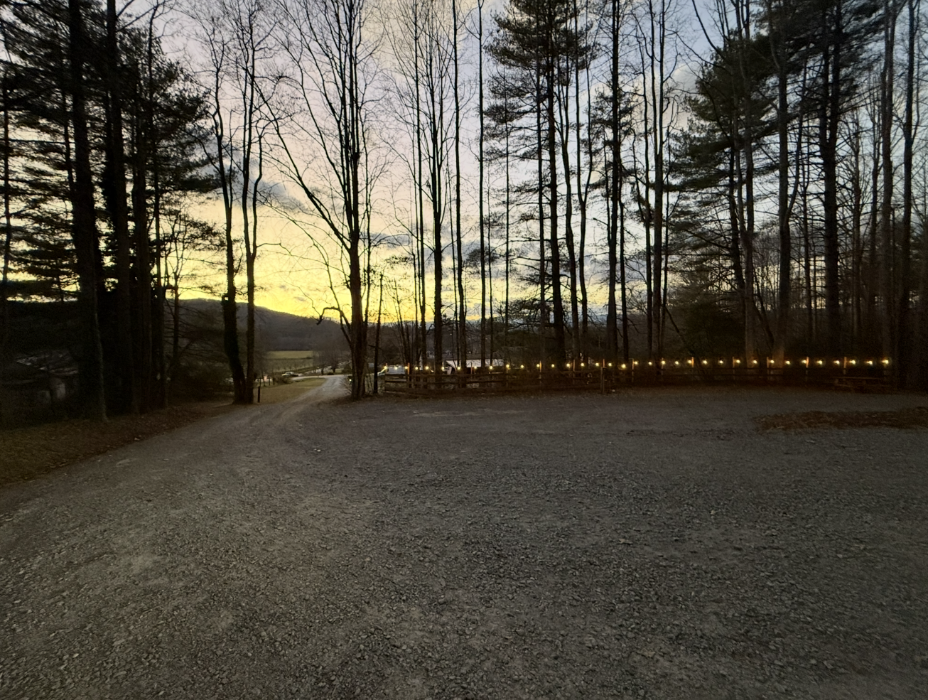 A gravel driveway in a wooded area at dusk, with leafless trees and a fence lined with lights. In the background, there are hills and a few vehicles parked along the road.