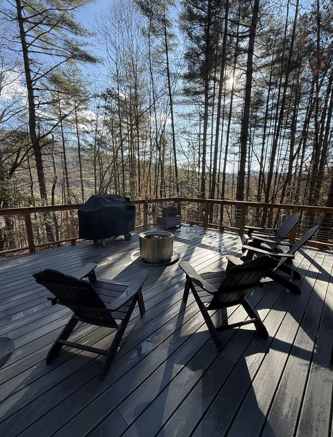 Wooden deck with outdoor chairs, a fire pit, a grill, and a basket of firewood, surrounded by tall trees and a clear blue sky.