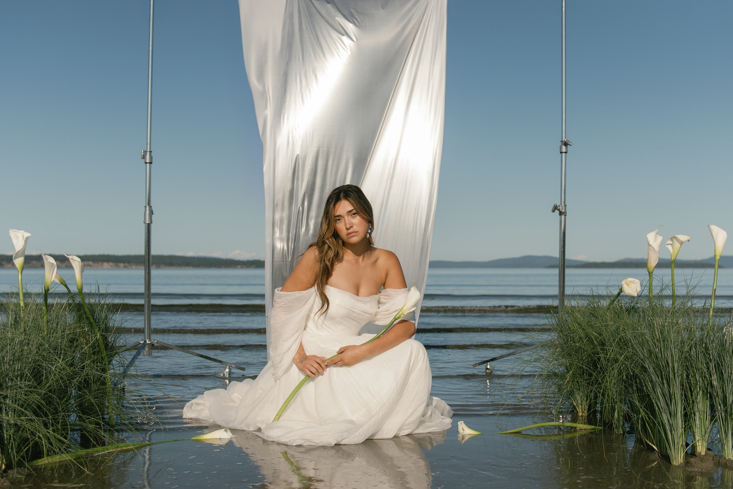 a stunning bride in a beautiful bridal gown posing near the water holding a calla lilly