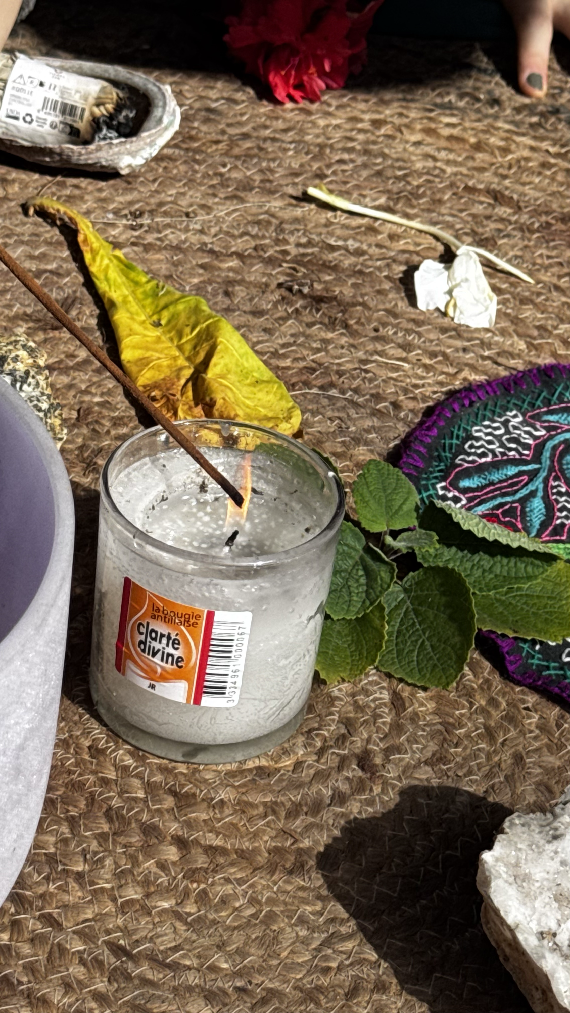 Cierge blanc avec une étiquette orange et blanche, entouré de feuilles vertes, placé sur un tapis en osier. L'allumette est allumée. Autres objets : une fleur rouge, un morceau de papier blanc, un tissu coloré, des papiers ou objets divers, et un fon