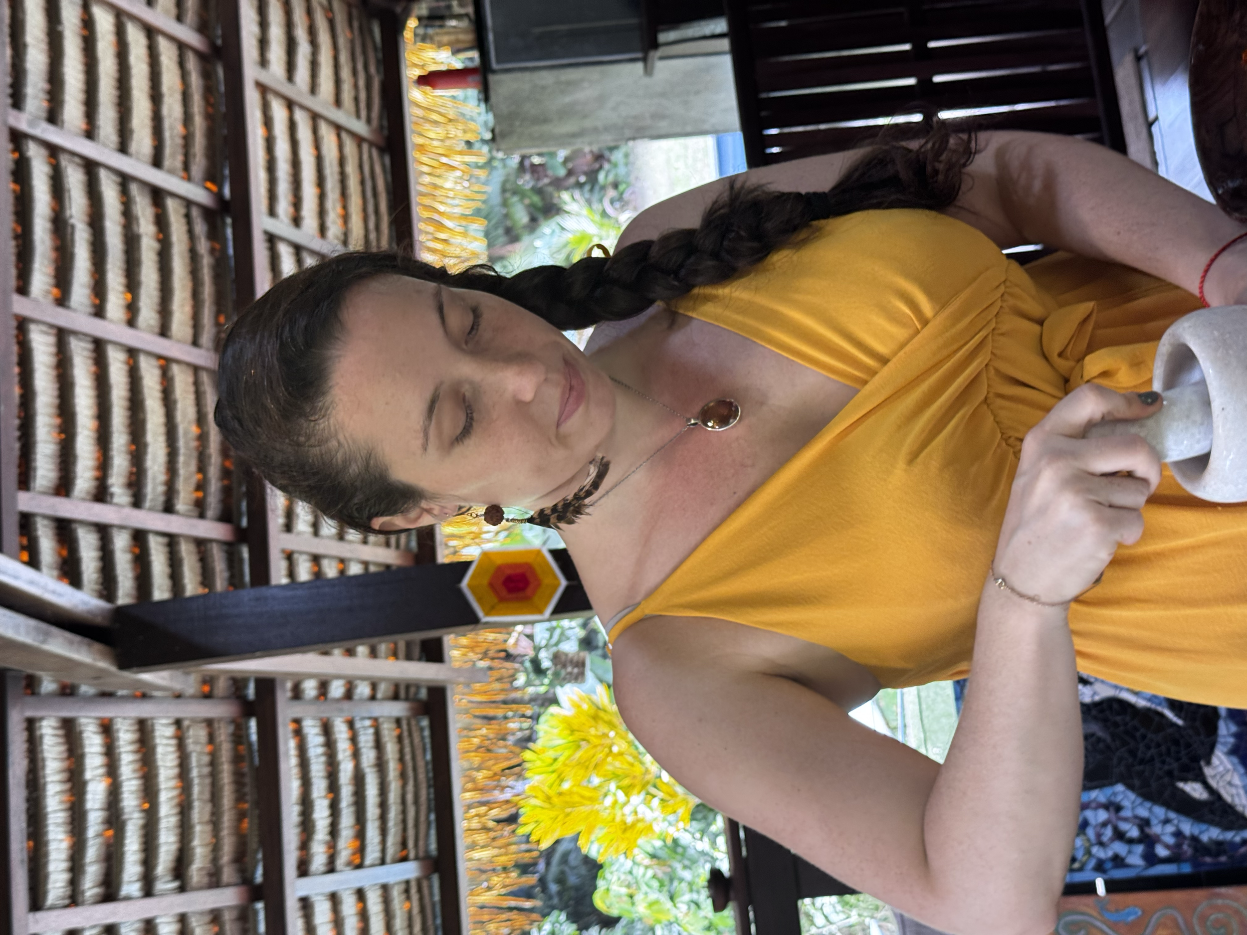 Jeune femme portant une robe jaune, assise à une table avec une tasse en main, décoration en arrière-plan avec fleurs jaunes et une structure en bois.