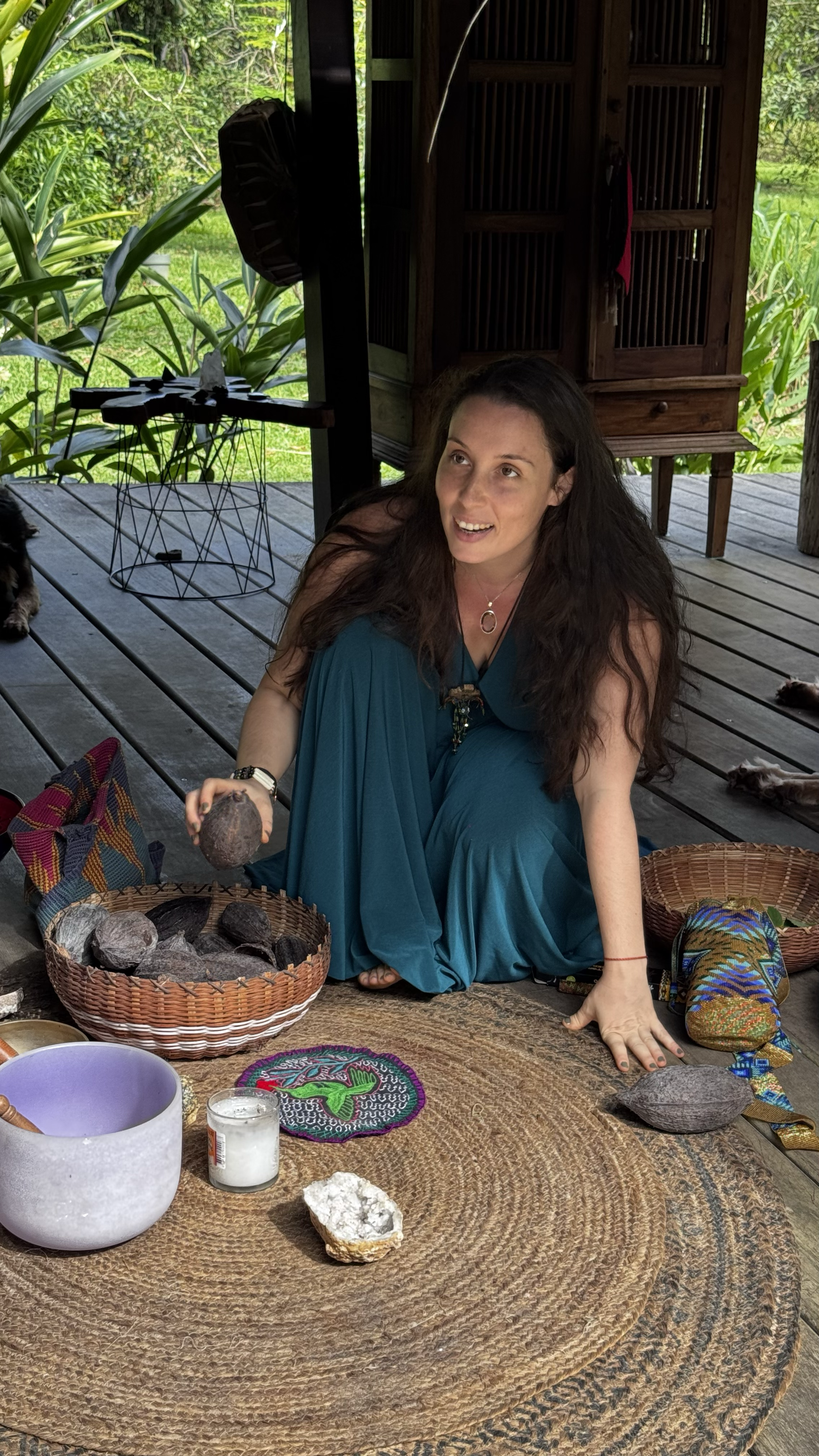 Une femme assise au sol en train de manipuler un gros caillou. Elle porte une robe bleue et est entourée d'autres objets comme des paniers, une bougie, et une coupe en pierre. L’arrière-plan montre une structure en bois avec des plantes vertes, évoqu