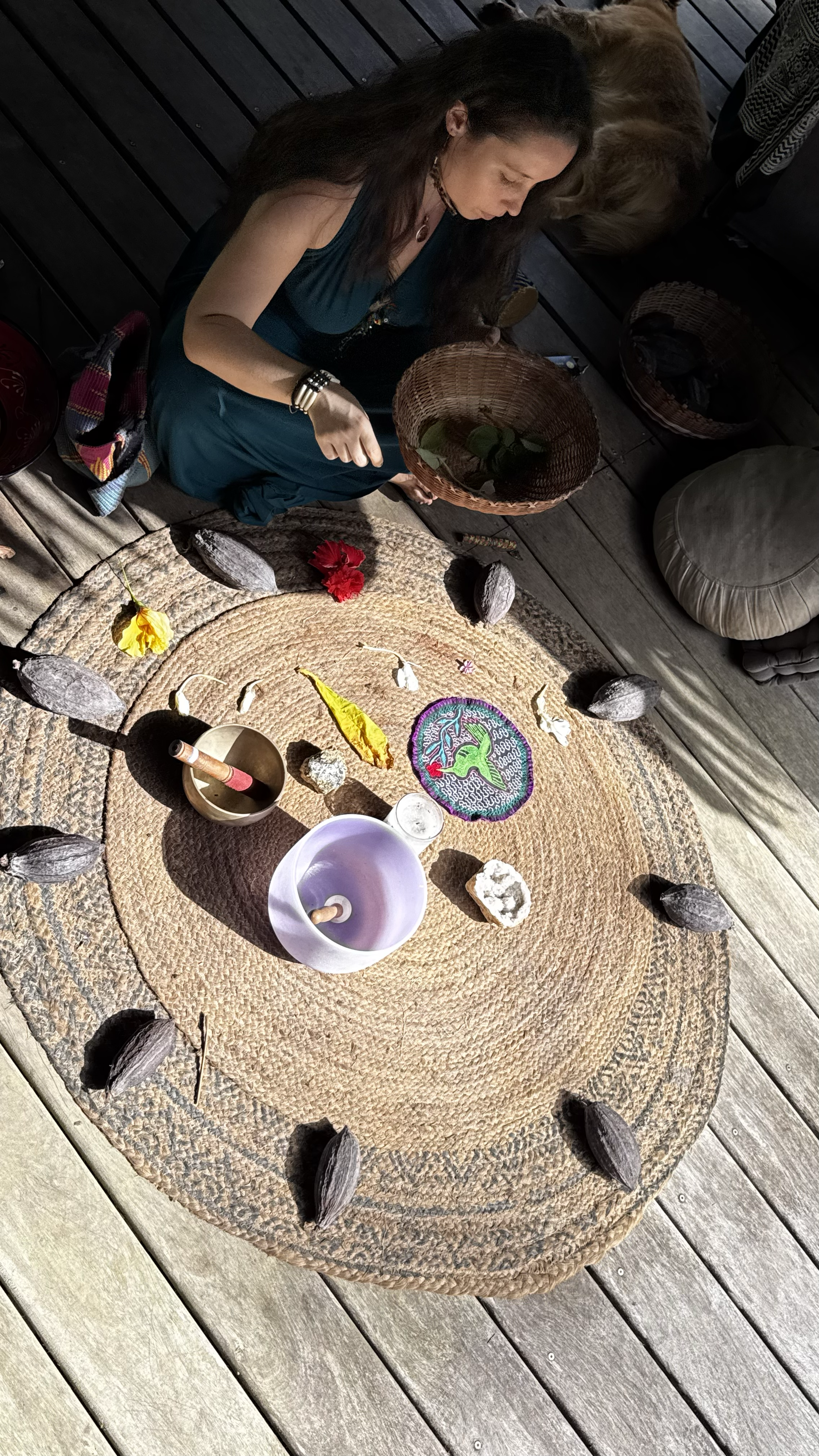 Une femme tenant un panier en osier, assise sur un tapis de fibres naturelles, entourée de pierres et de fleurs, dans une ambiance de méditation ou de rituel spirituel.